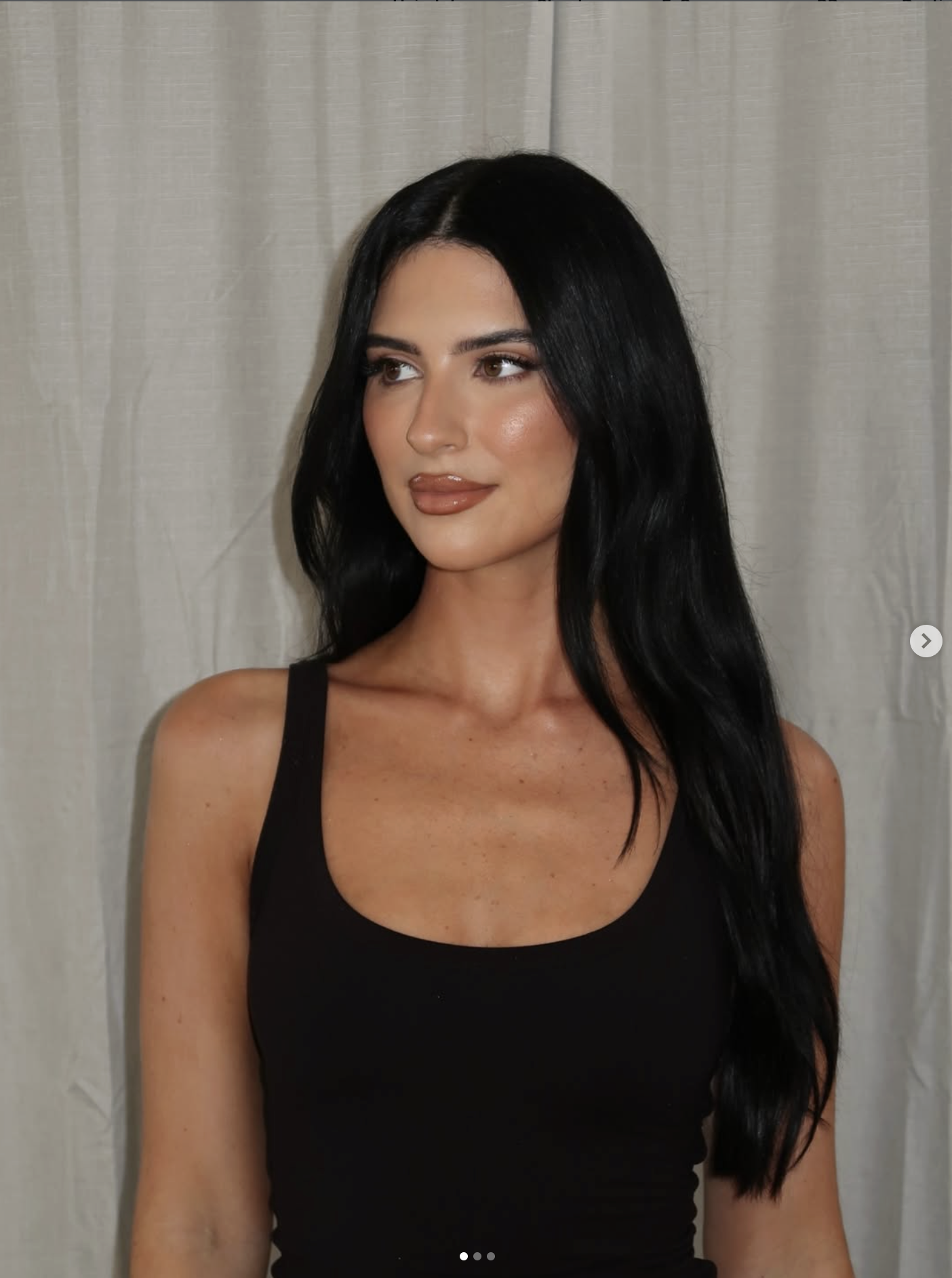 Portrait of a woman with long black hair wearing a black sleeveless top, standing against a light-colored curtain background, looking to her left.