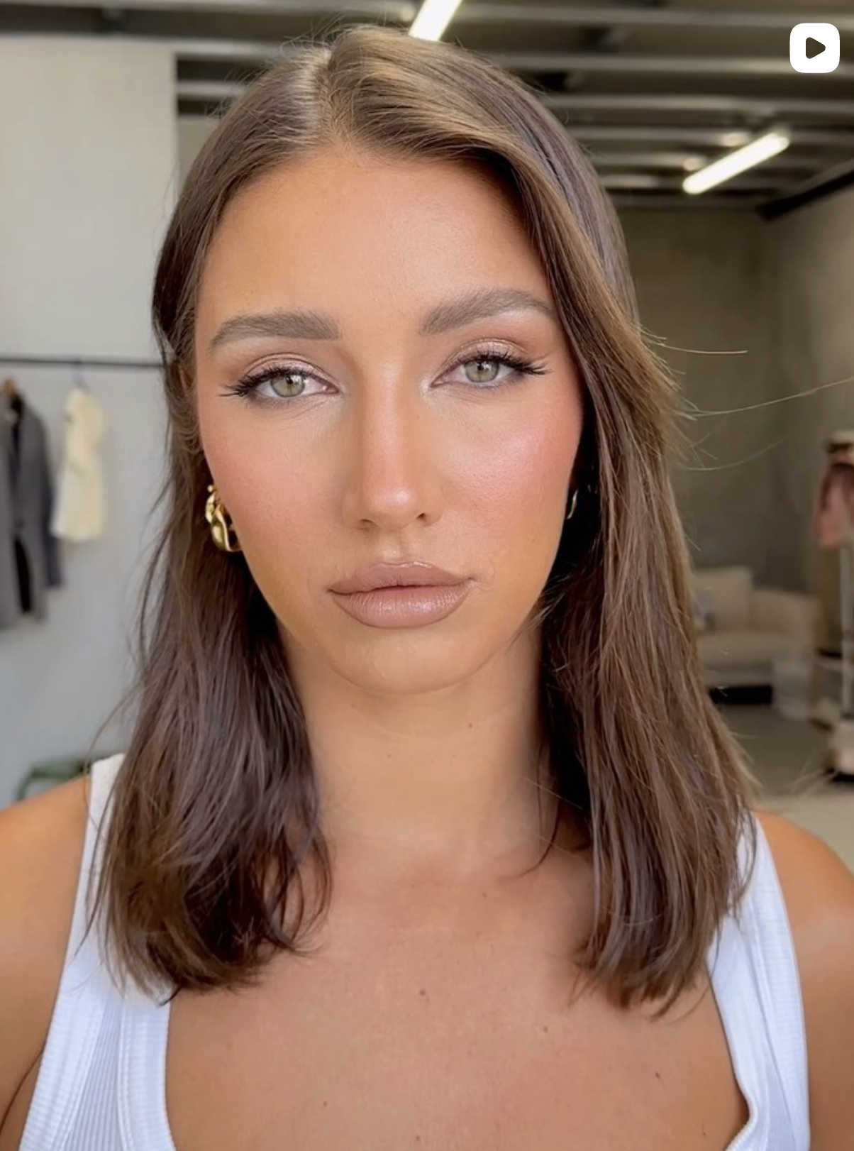 Close-up of a woman with long brown hair, green eyes, and makeup, wearing a white tank top and gold hoop earrings, standing in an indoor space with clothing and people in the background.