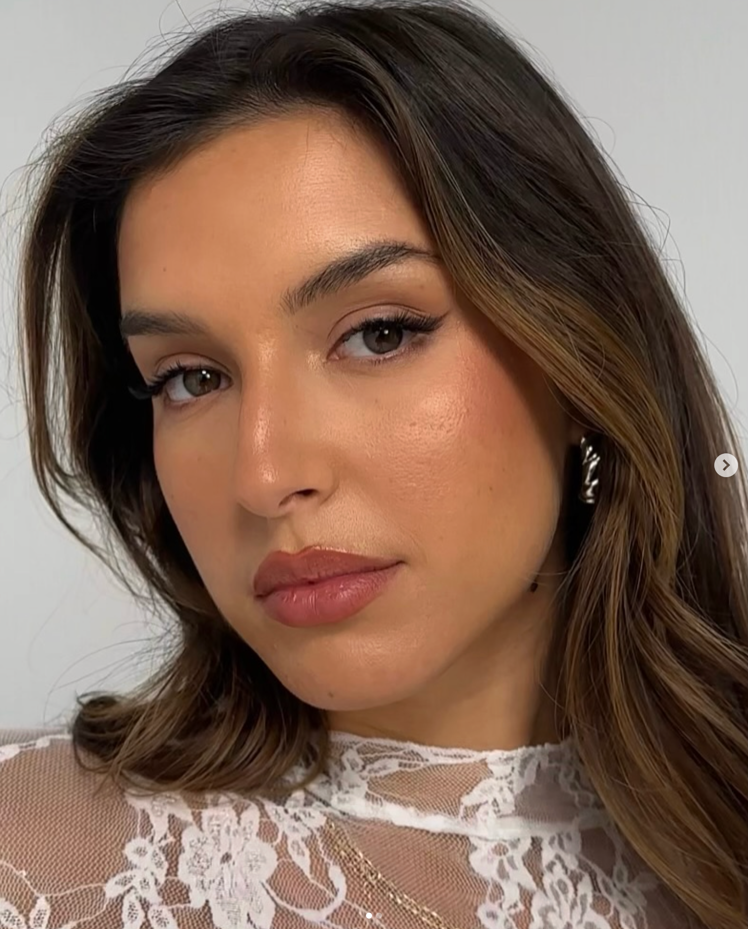 Close-up of a woman with medium-length brown hair styled in loose waves, wearing a lace top, with makeup including eyeliner, mascara, and pink lipstick, against a plain background.