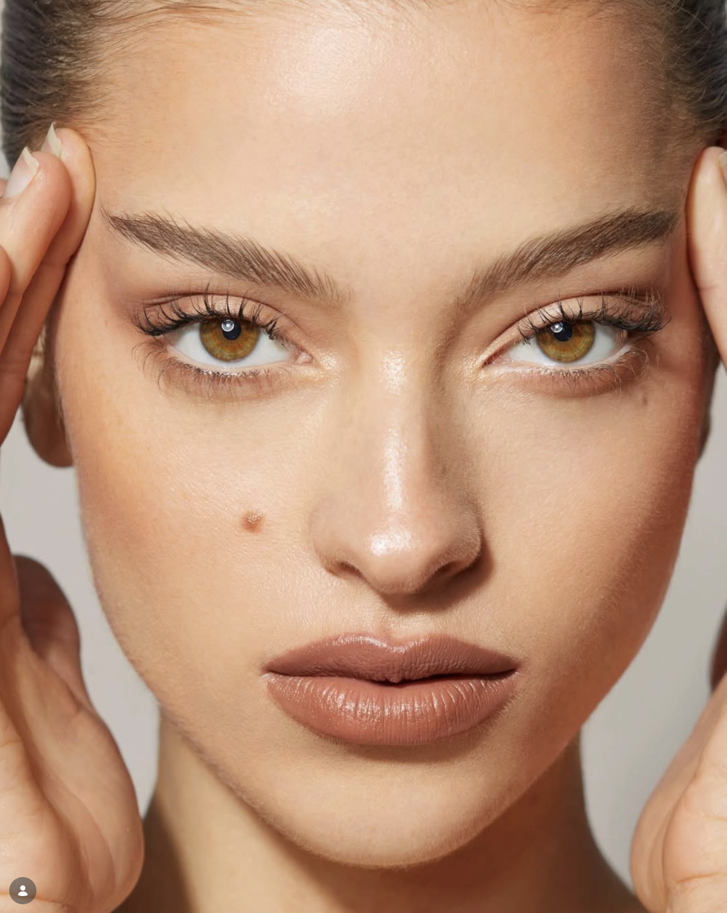Close-up of a woman's face with brown eyes, full lips, groomed eyebrows, and smooth skin, touching her temples with her hands.