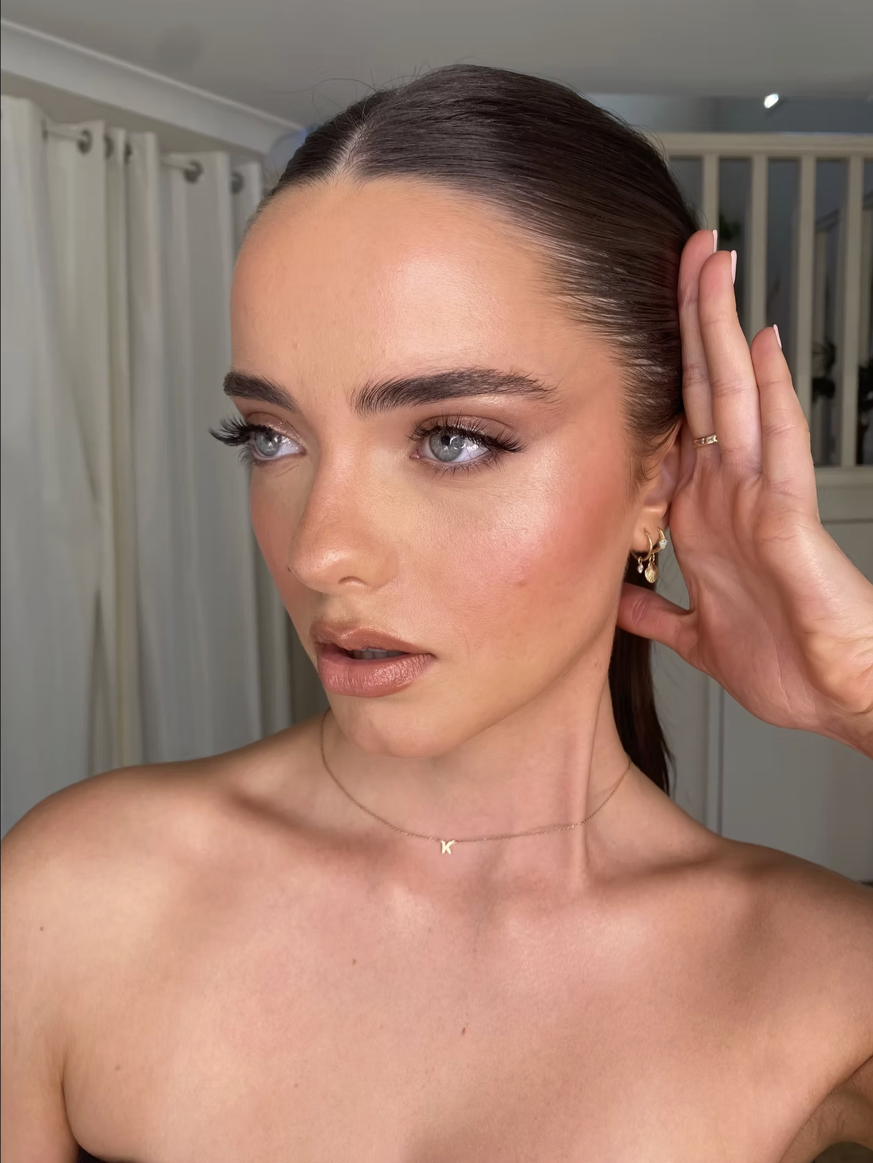 Close-up of a woman with dark brown hair, blue eyes, and makeup, touching her ear and looking slightly to the side. She is wearing jewelry including earrings, a ring, and a necklace with a letter 'K'.
