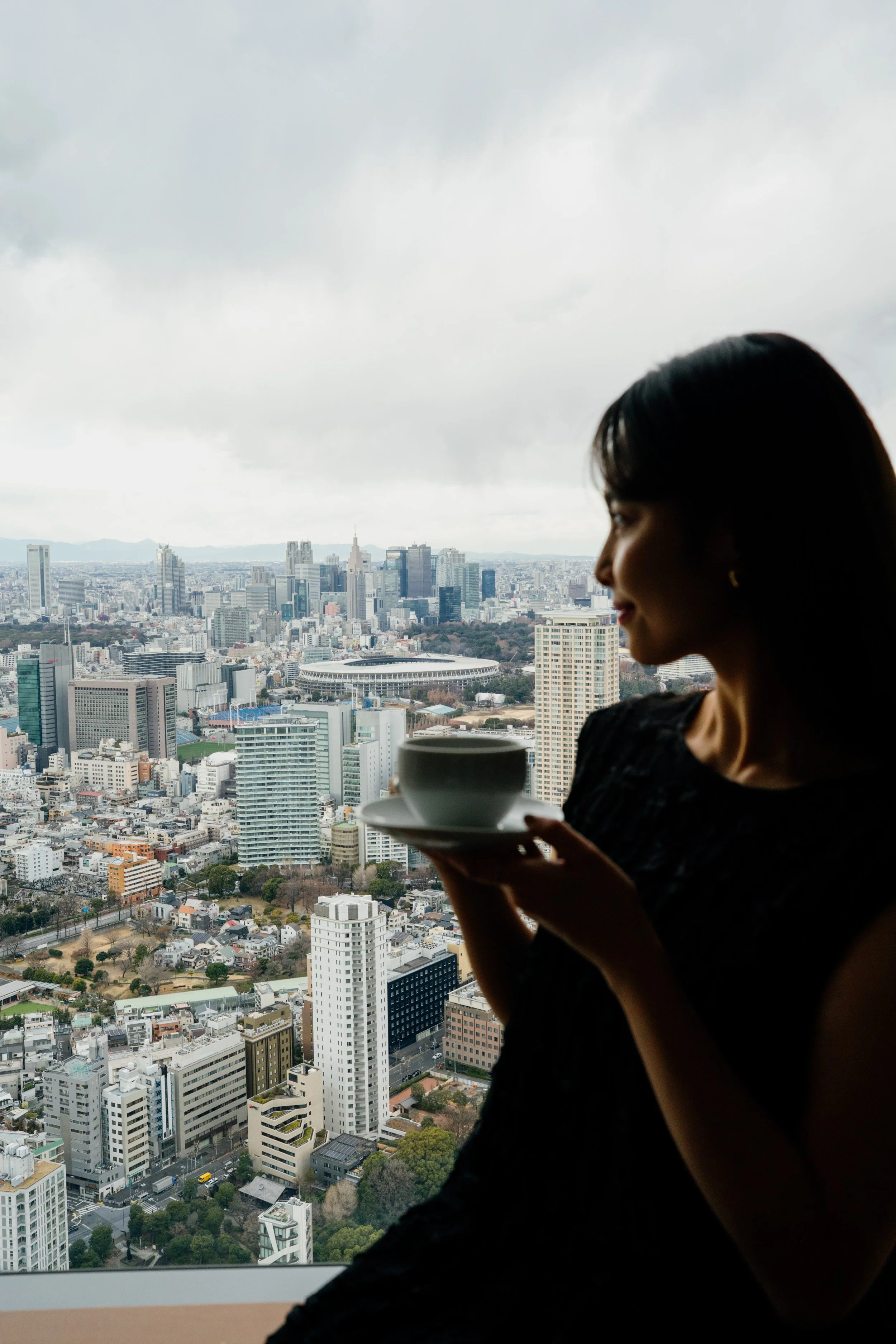 A woman with dark hair holding a white cup and saucer, with a city skyline and cloudy sky in the background at The Ritz-Carlton, Tokyo. Editorial hotel photography, Japan.