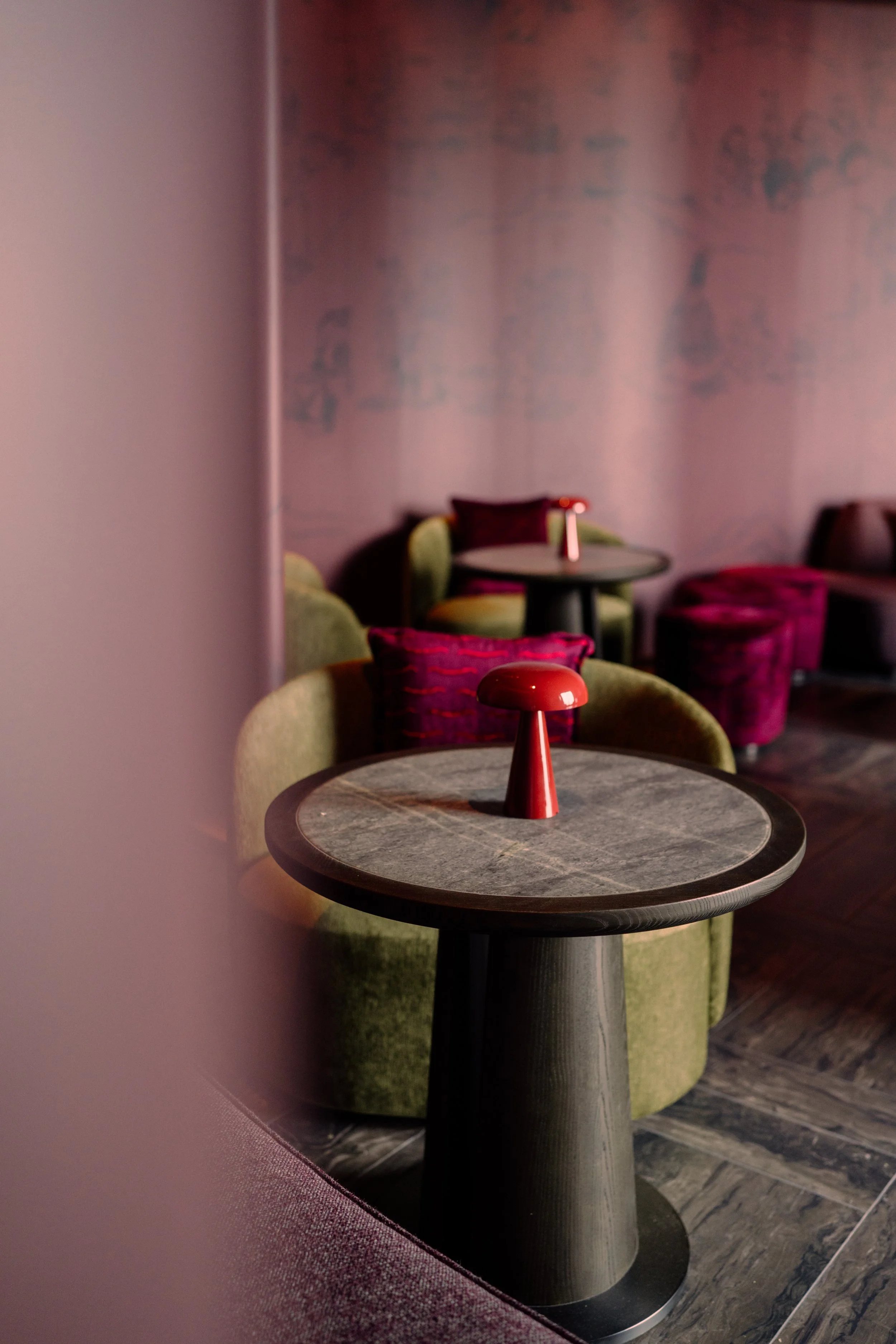 Empty tables with colorful velvet chairs in a dimly lit lounge or bar setting, with a pink wall and abstract artwork in the background.