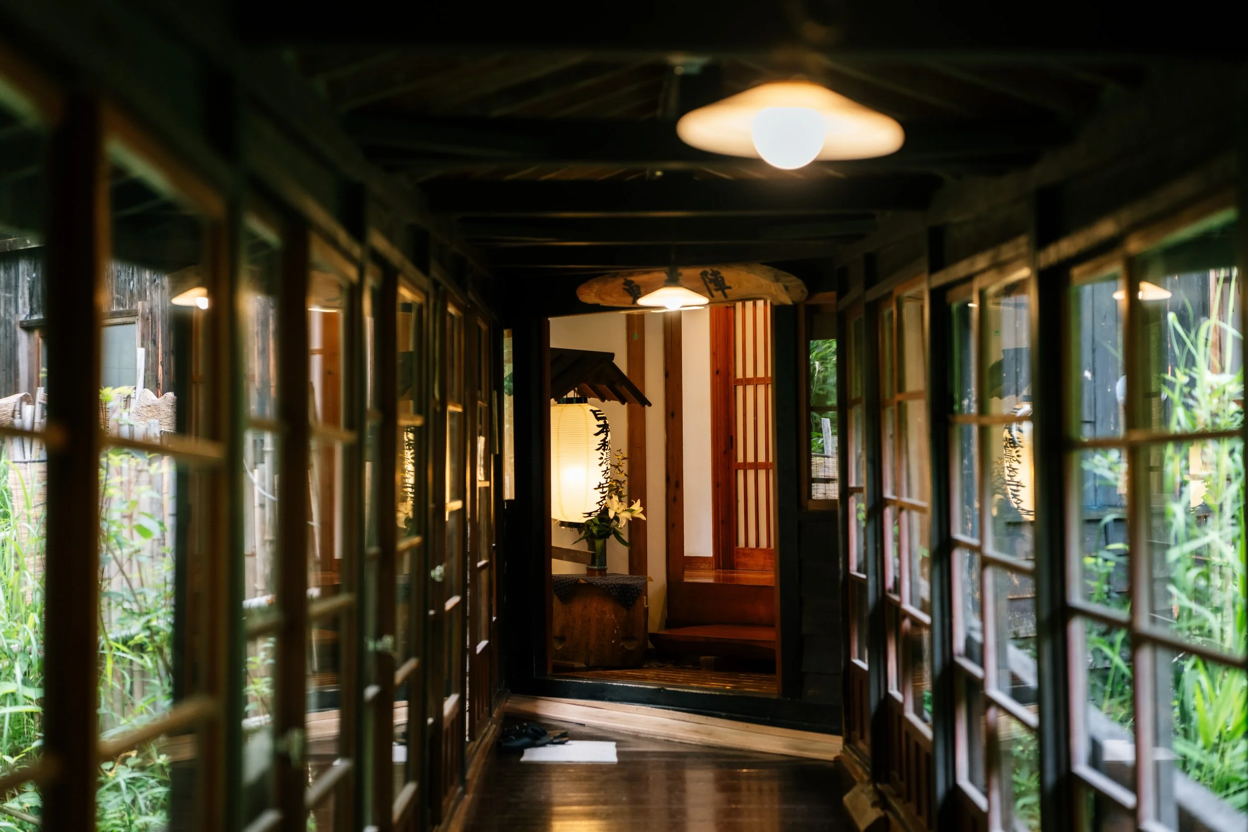 View of traditional Japanese-style interior corridor with wooden frames and shoji doors, leading to a room with Japanese lantern and flowers, with greenery outside.