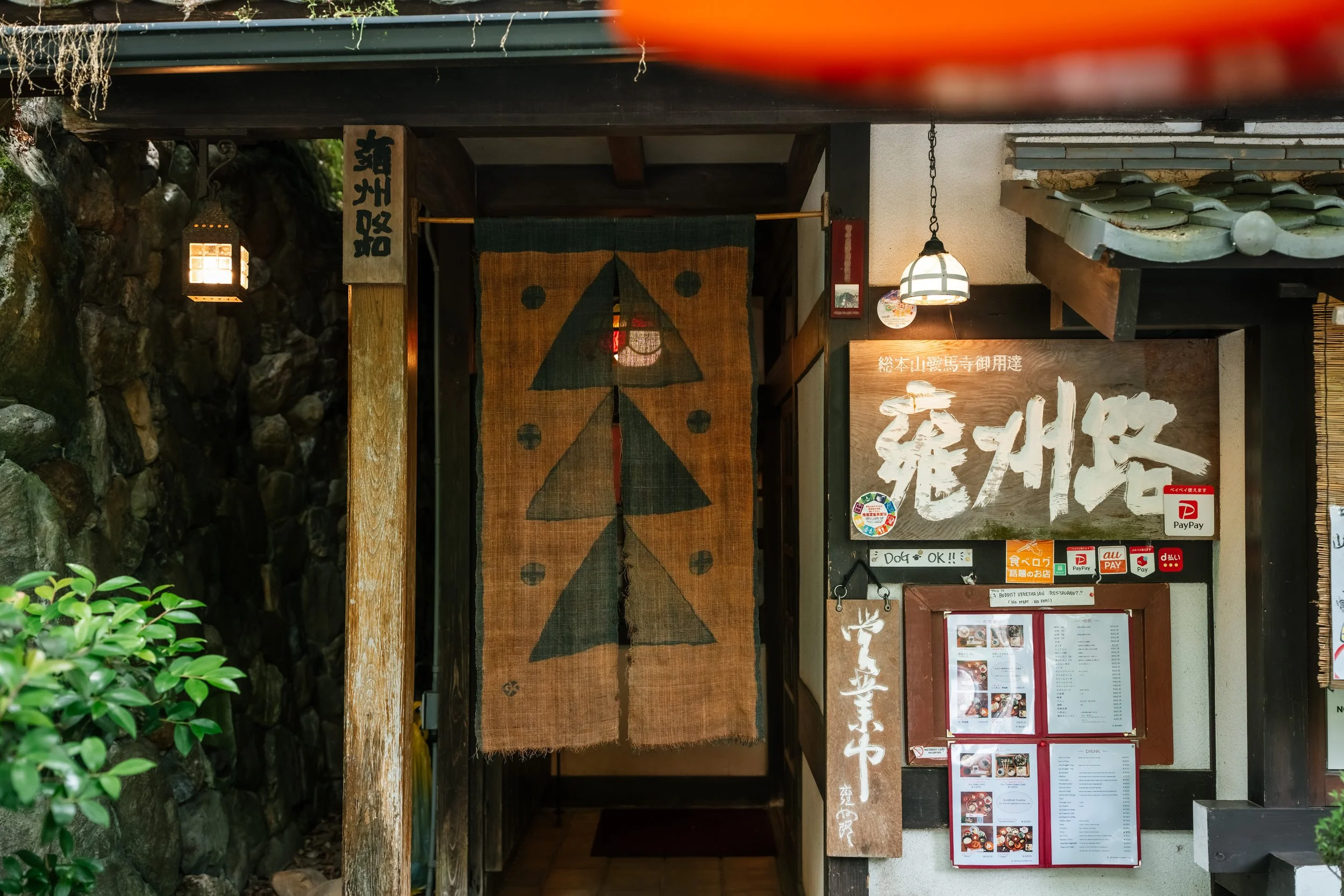 Entrance to a Japanese restaurant or shop with a fabric curtain featuring a tree symbol and Japanese characters, lantern lights, and a menu display outside.