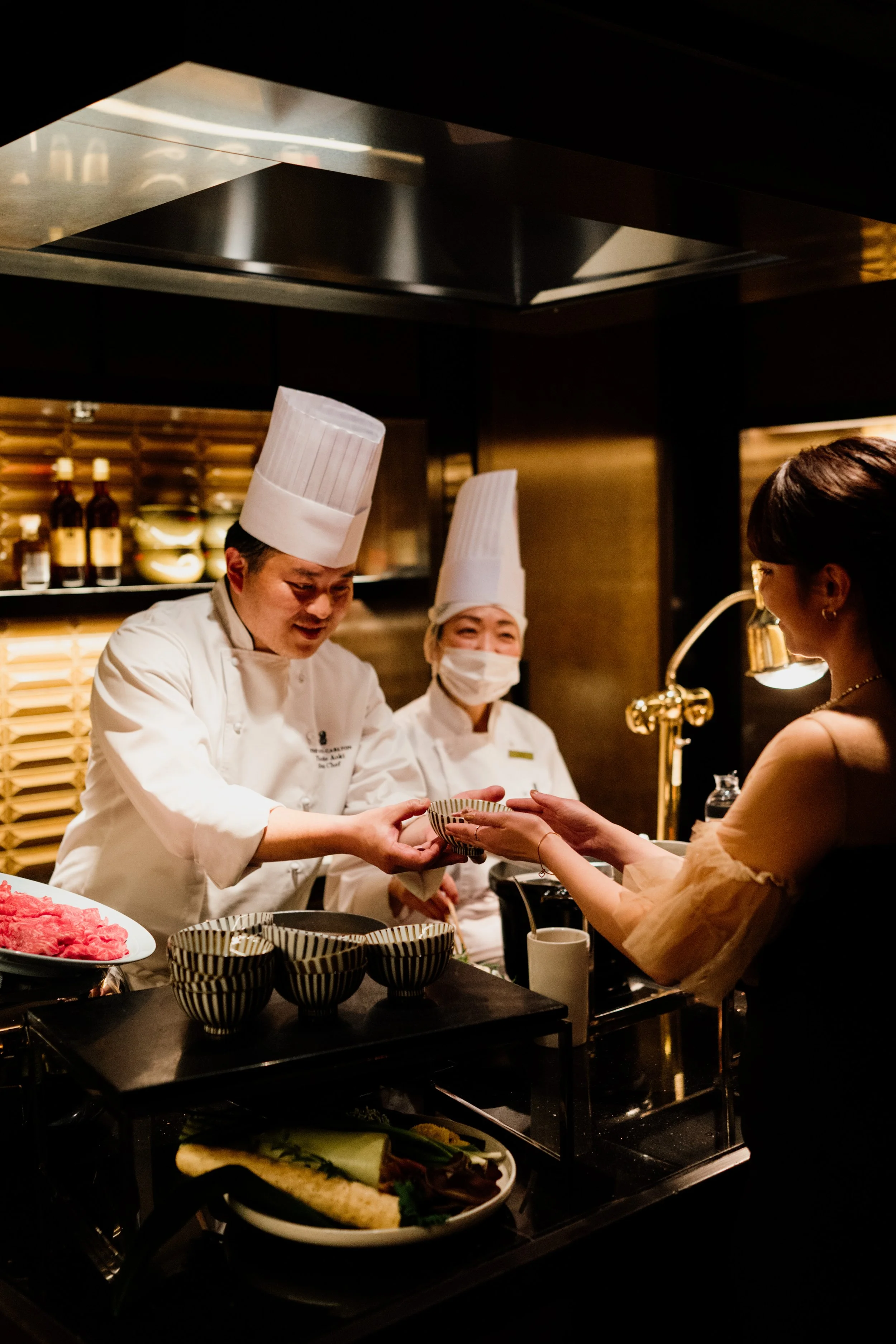 Chefs in white uniforms and tall hats serve food to a woman at a buffet restaurant, with dark interior and warm lighting at The Ritz-Carlton, Tokyo. Editorial hotel photography, Japan.