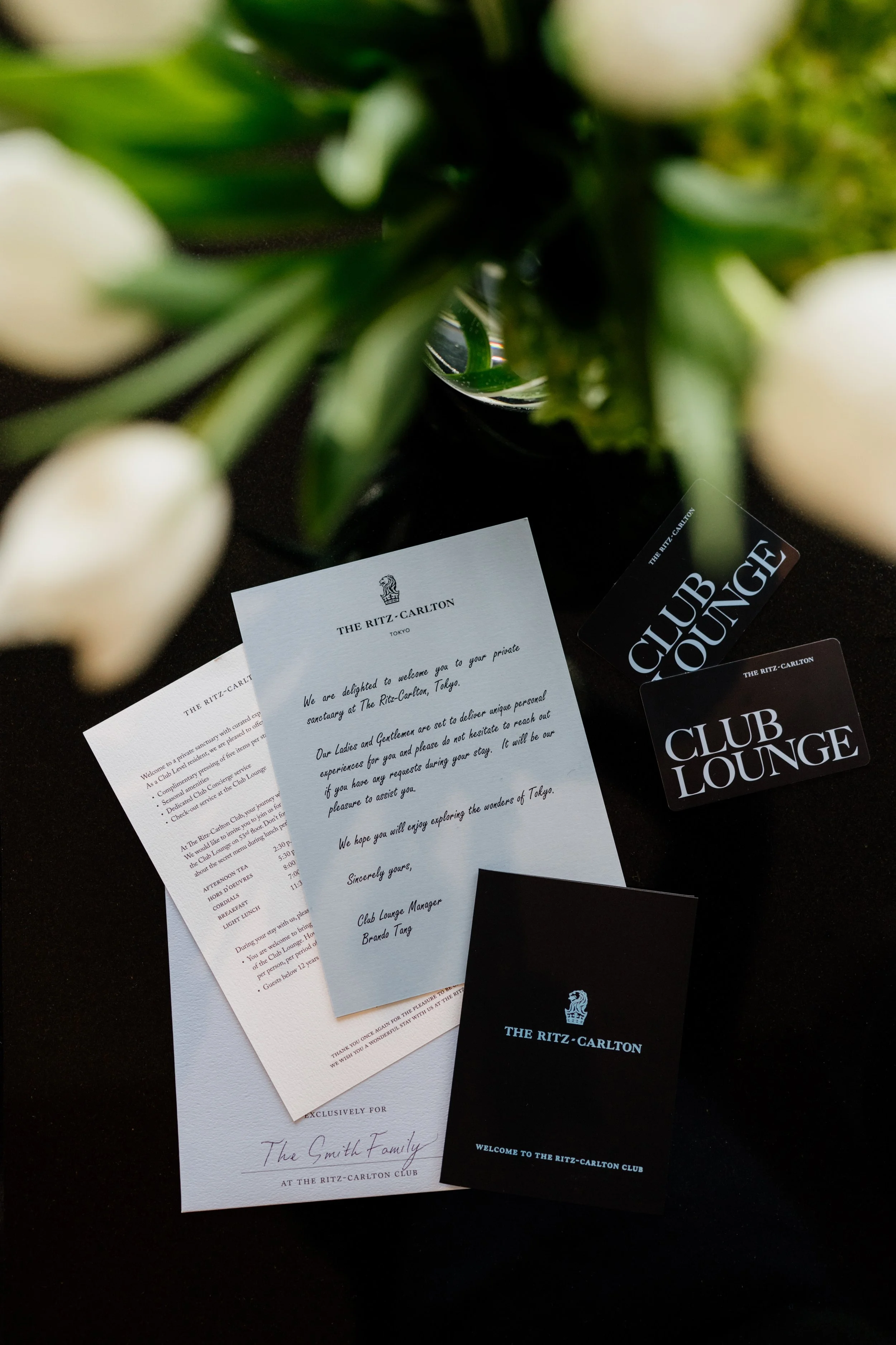 Invitation letter and club cards from The Ritz-Carlton, Tokyo, placed on a dark surface, partially obscured by a bouquet of white flowers, editorial hotel photography, Japan.