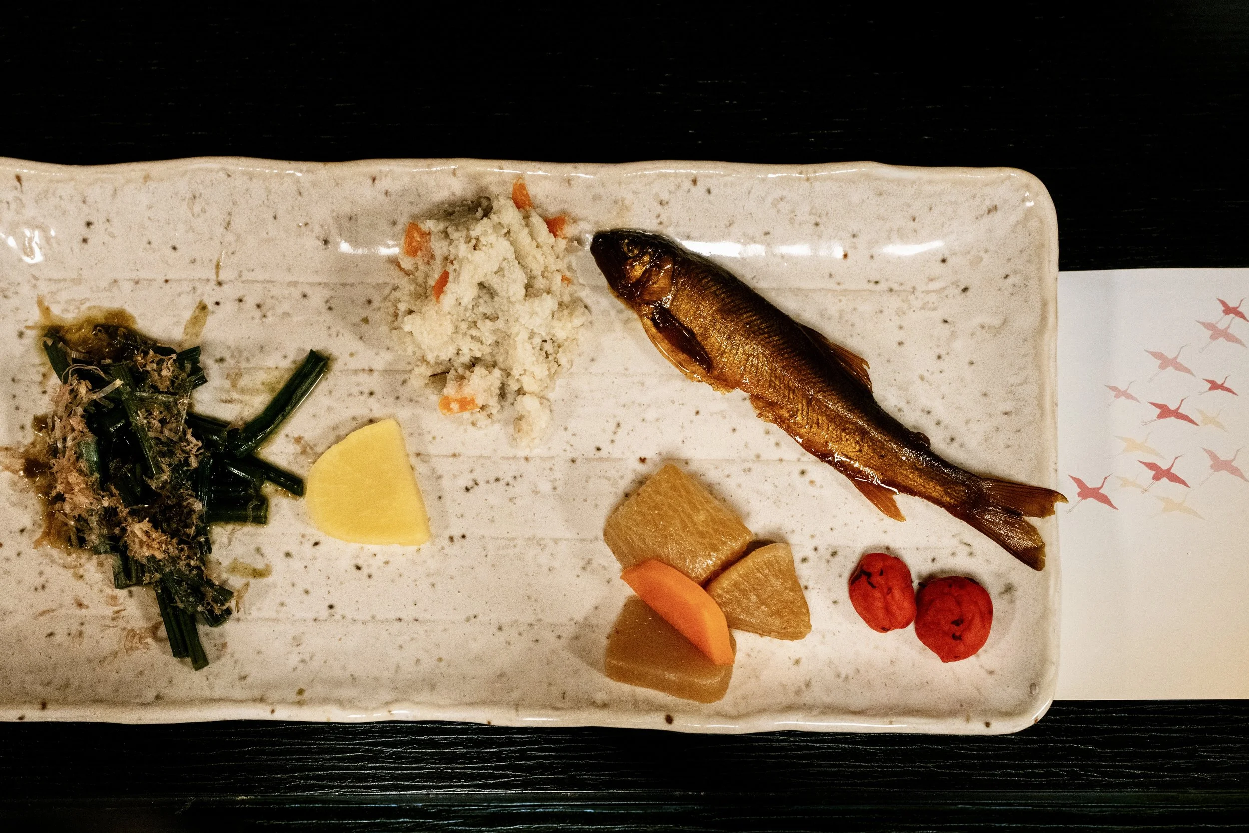 A rectangular ceramic plate with various types of food, including grilled fish, mashed potatoes with carrots, cooked greens, sliced lemon, and small red fruit or berry, on a black surface.