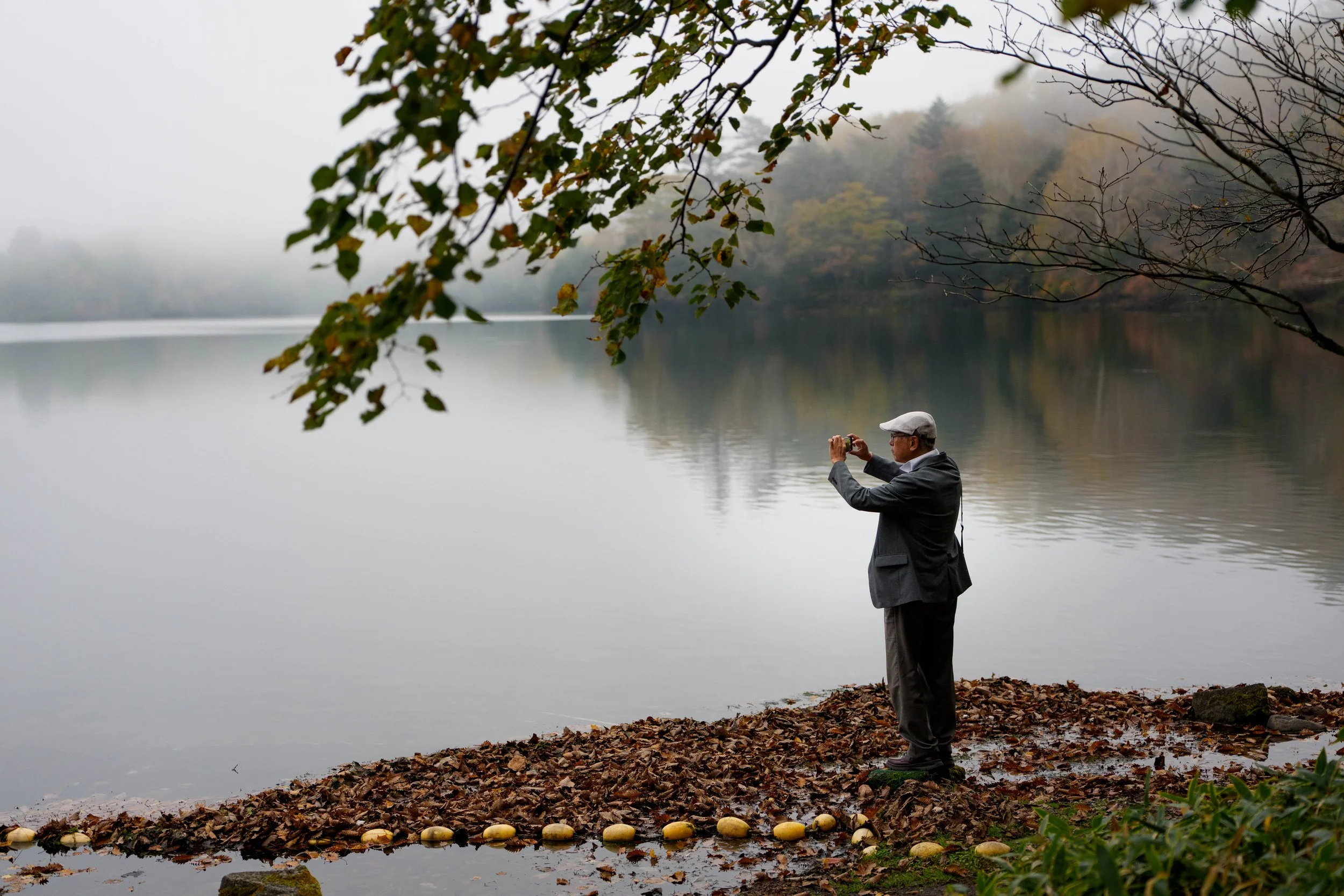 An elderly man standing on a leaf-covered shore of Lake Yuno in Oku-Nikko with overhanging tree branches and autumn-coloured leaves frame the scene, travel photography in Japan.