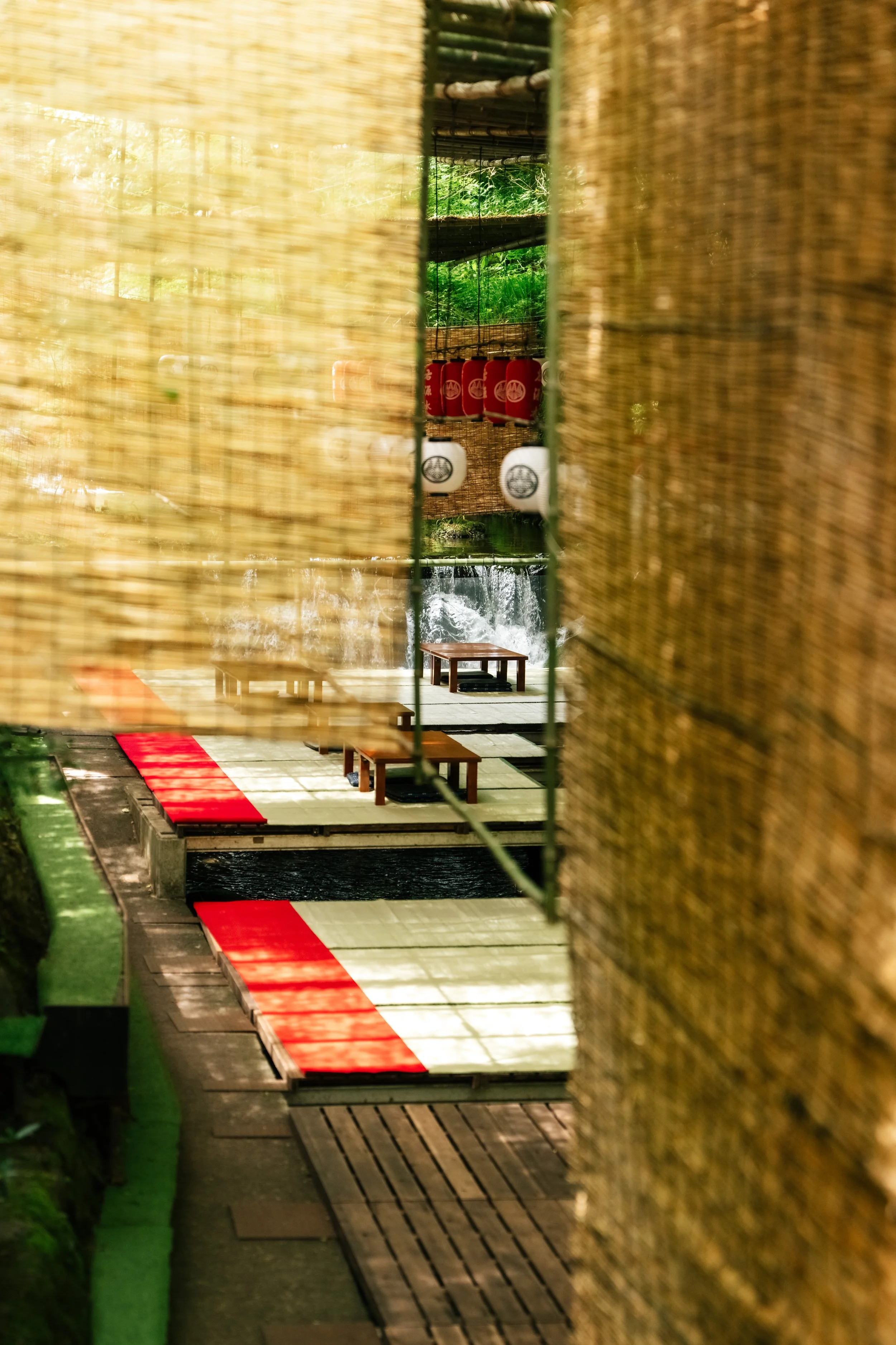 A view through a narrow space showing a Japanese-inspired outdoor setting with lanterns, a wooden platform, and decorative elements, framed by bamboo or reed fencing.