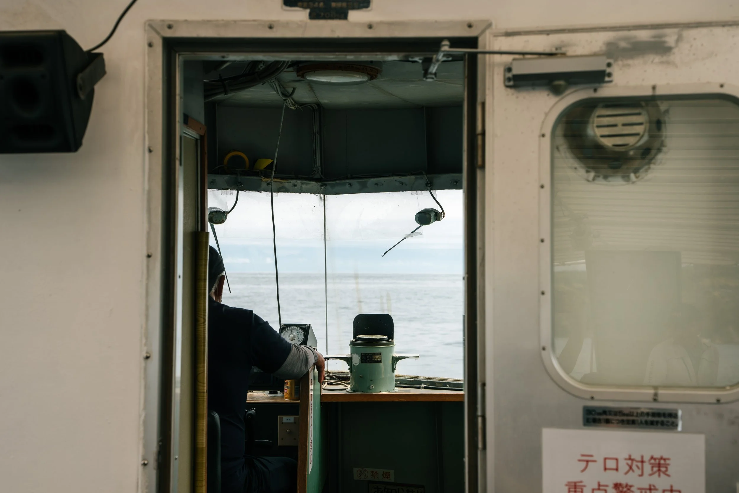 View from inside a boat showing a person operating navigational equipment, looking out over the ocean through an open window or portal. Noto, Ishikawa, editorial travel photography, Japan.