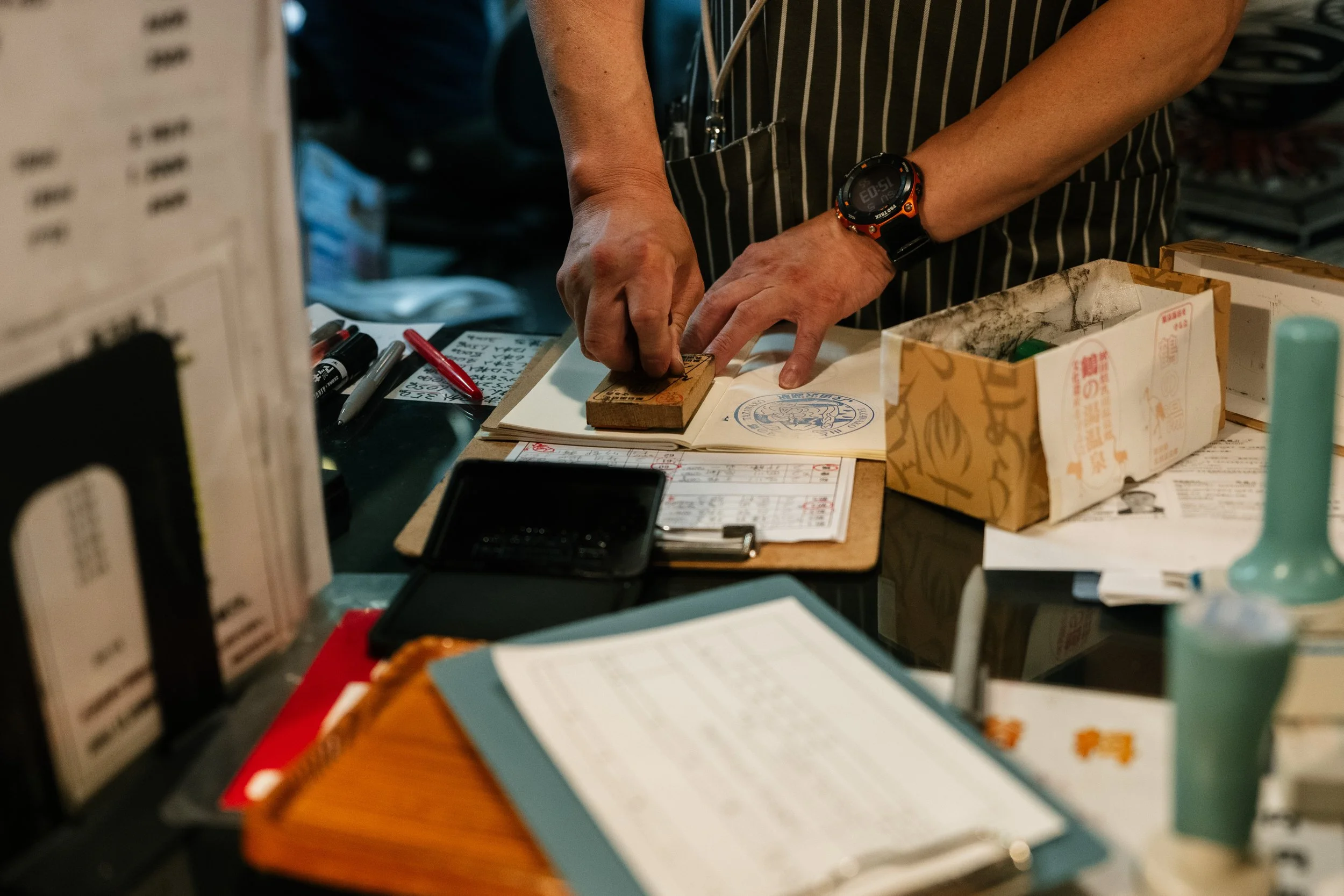Person's hands stamping a document at a checkout counter. The person is wearing a black and orange watch and a striped apron. Various papers, a pen, and a cardboard box are visible on the counter.