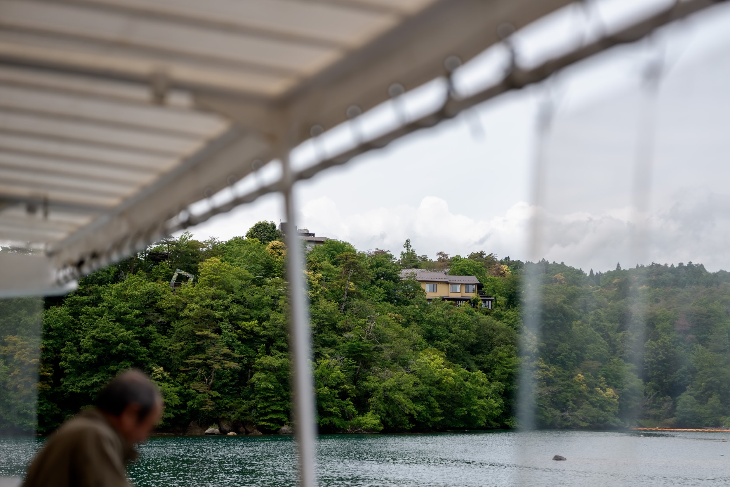 A lake surrounded by lush green trees with houses on a hillside in the background, viewed from underneath a white boat canopy, with a person's blurred head in the foreground.