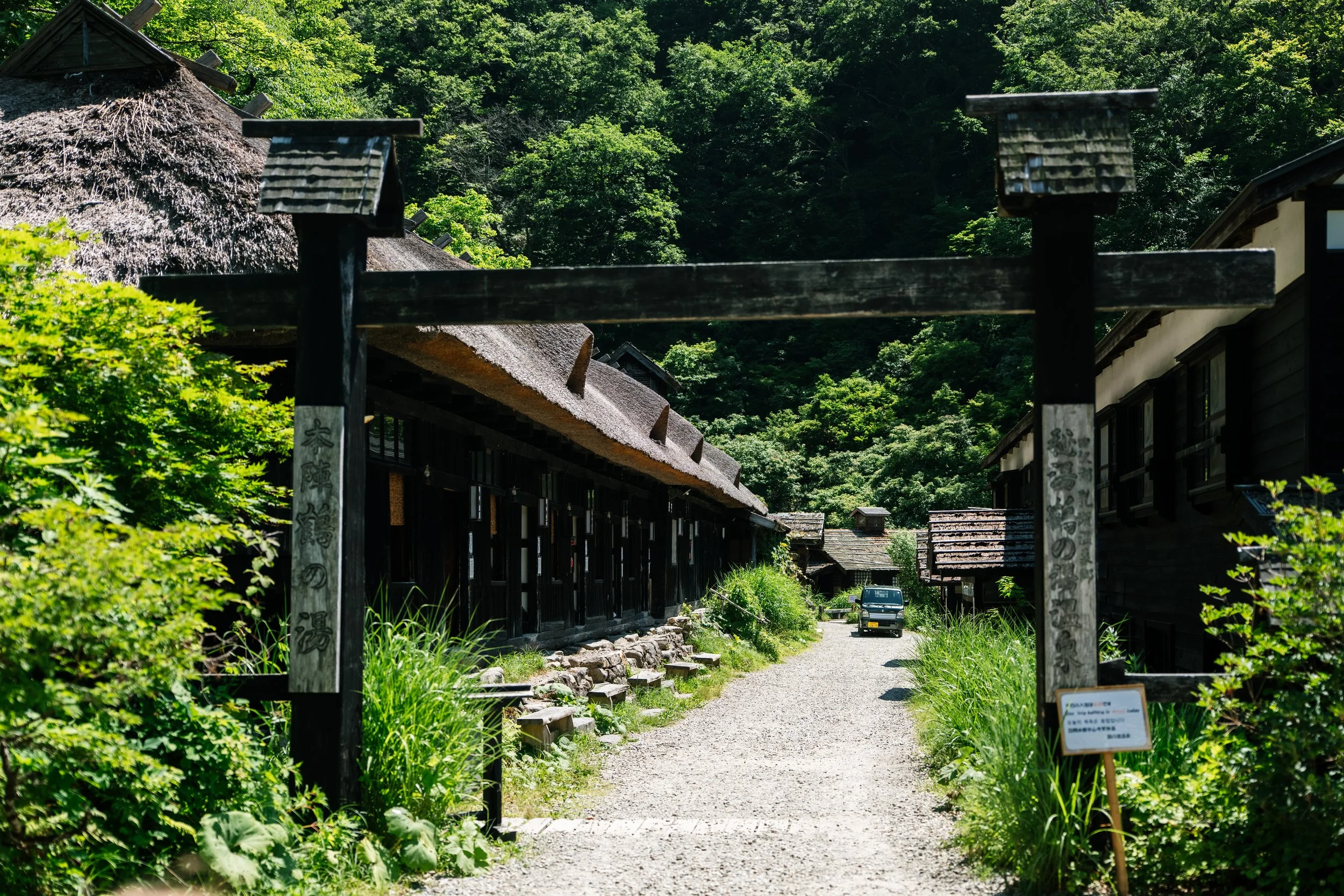 A pathway through a traditional village with wooden buildings, surrounded by lush green trees, and a vehicle parked at the end of the path. There is a wooden gate at the entrance with Japanese characters on the posts.