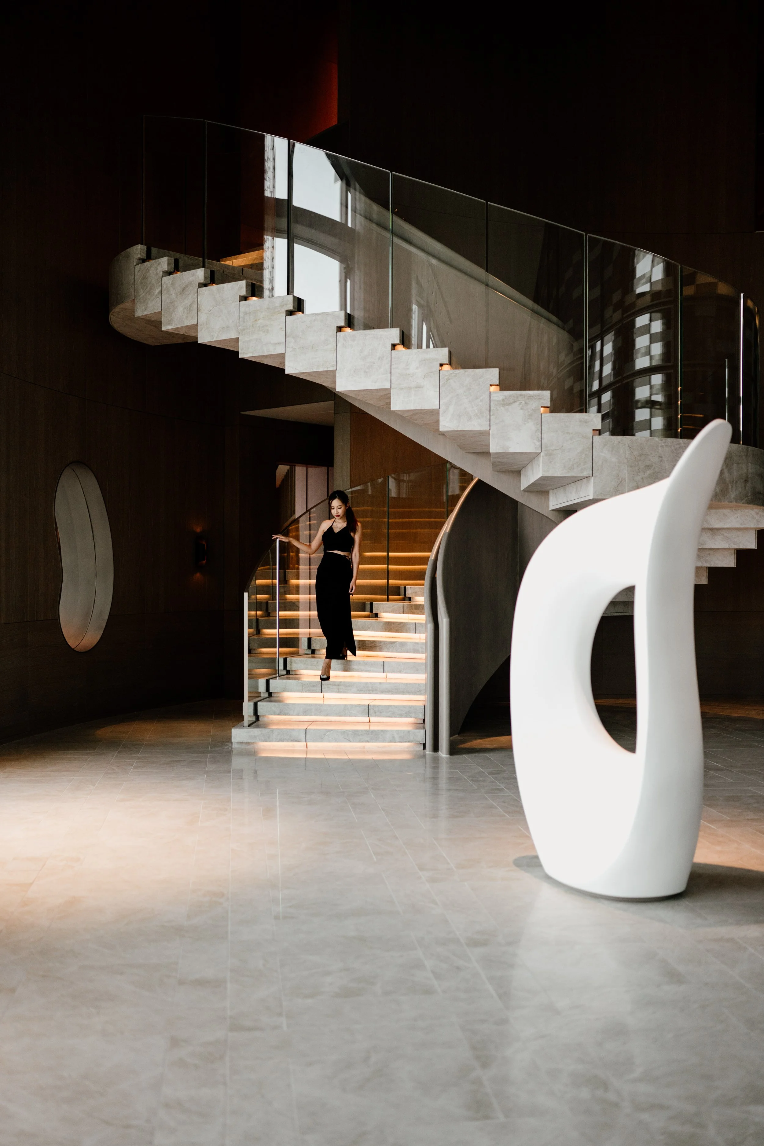 Modern hotel interior with a marble staircase, a woman in a black dress walking, a large white sculpture in the foreground, dark walls, and large windows with reflections at JW Marriott Tokyo hotel, editorial hospitality photography, Japan.