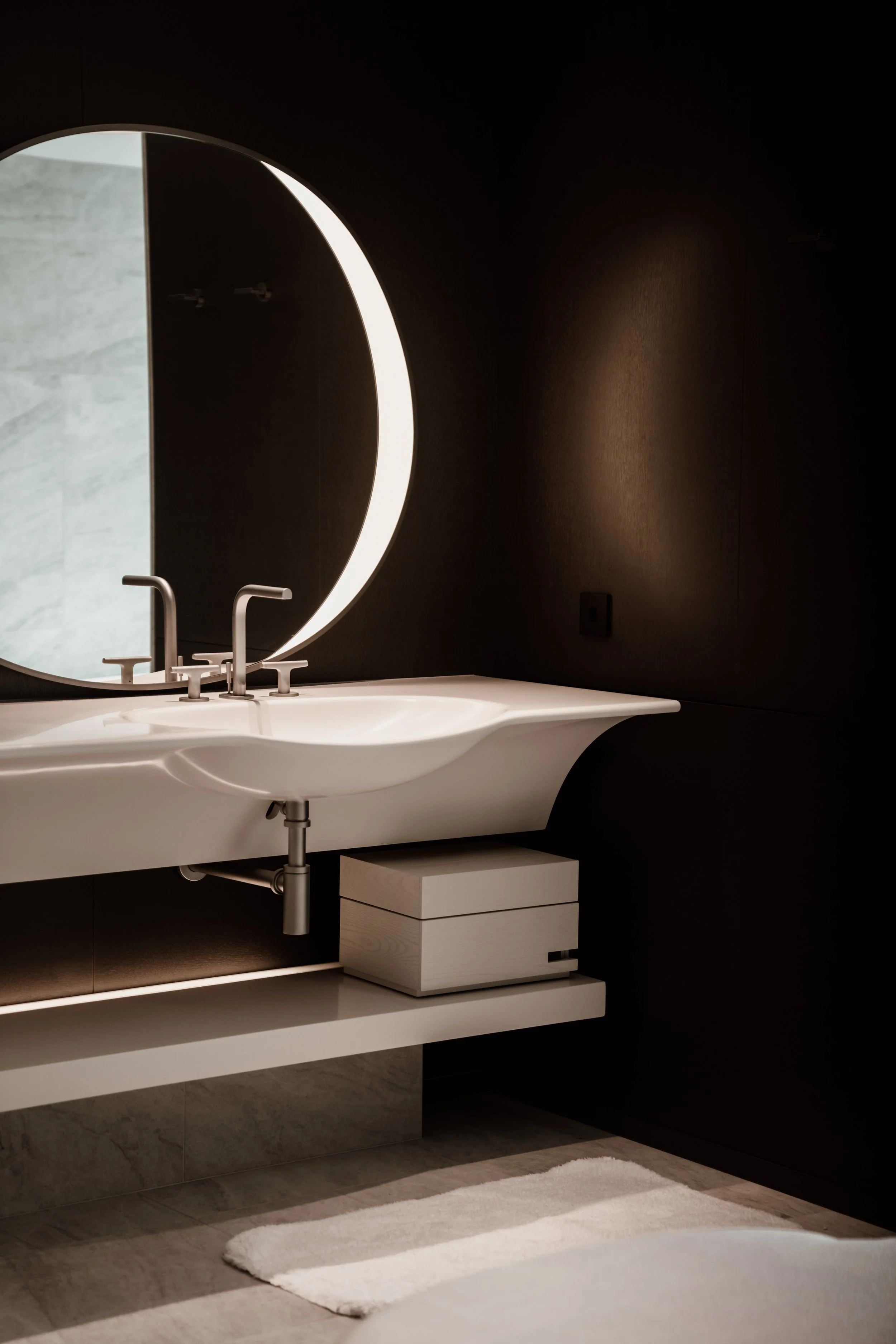 Modern bathroom with a white countertop sink, round mirror with built-in lighting, dark walls, light-colored floor, small wooden drawer, and a white rug at JW Marriott hotel, editorial hotel photography, Japan.