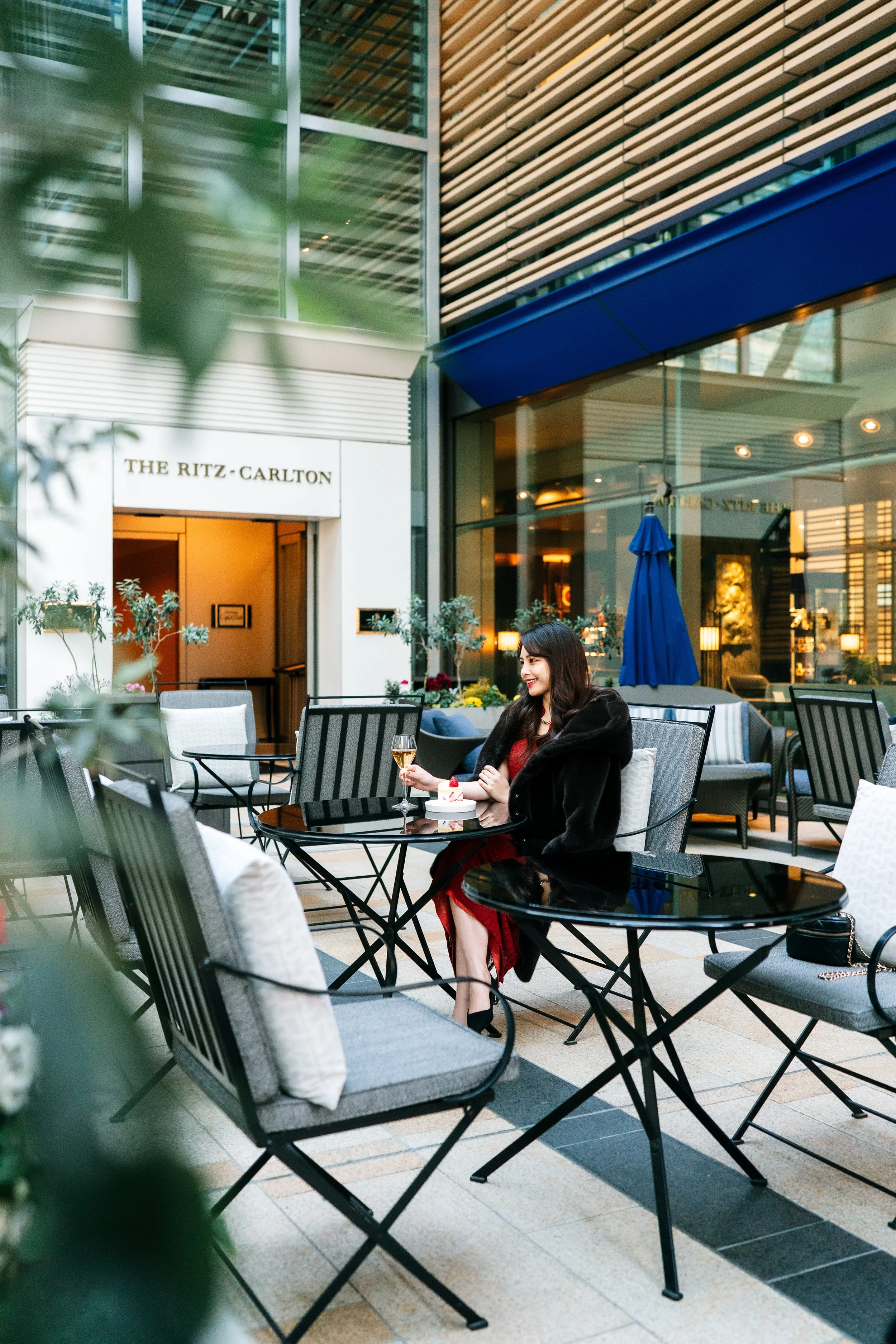 A woman in a red dress and black coat sitting at an outdoor cafe table, holding a glass of white wine and a plate with a dessert, in front of a glass building with the sign 'The Ritz-Carlton'. Editorial hotel photography, Japan.