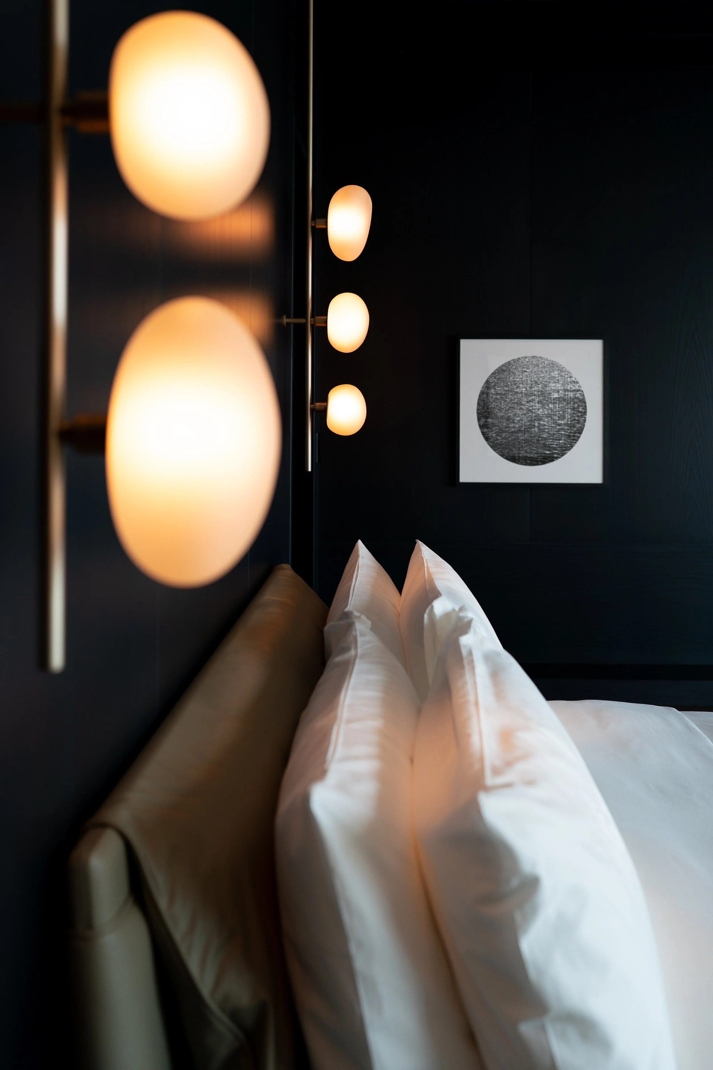 Close-up of a beige headboard with white pillows on a bed, modern wall-mounted lamps with oval-shaped frosted glass shades, dark wall with a framed black and white circular artwork.
