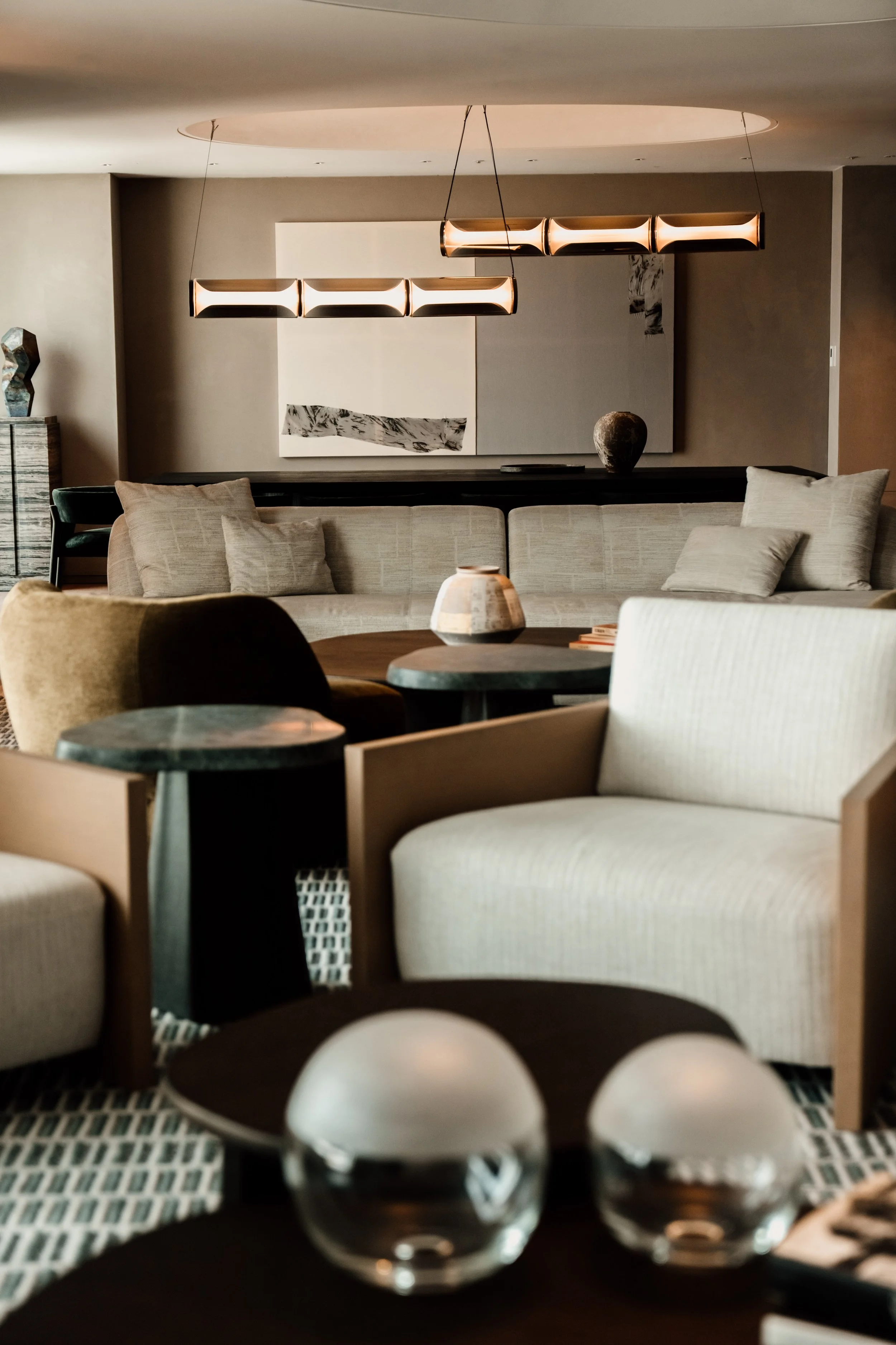 Modern living room with beige sofa, abstract wall art, contemporary hanging light fixture, decorative vase, and a patterned carpet.