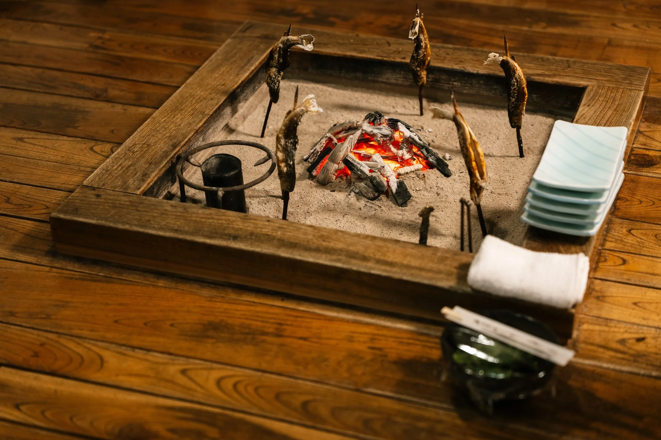 A tabletop grill with burning charcoal and skewered fish cooking over the embers, surrounded by a wooden frame and stacked white plates to the side.