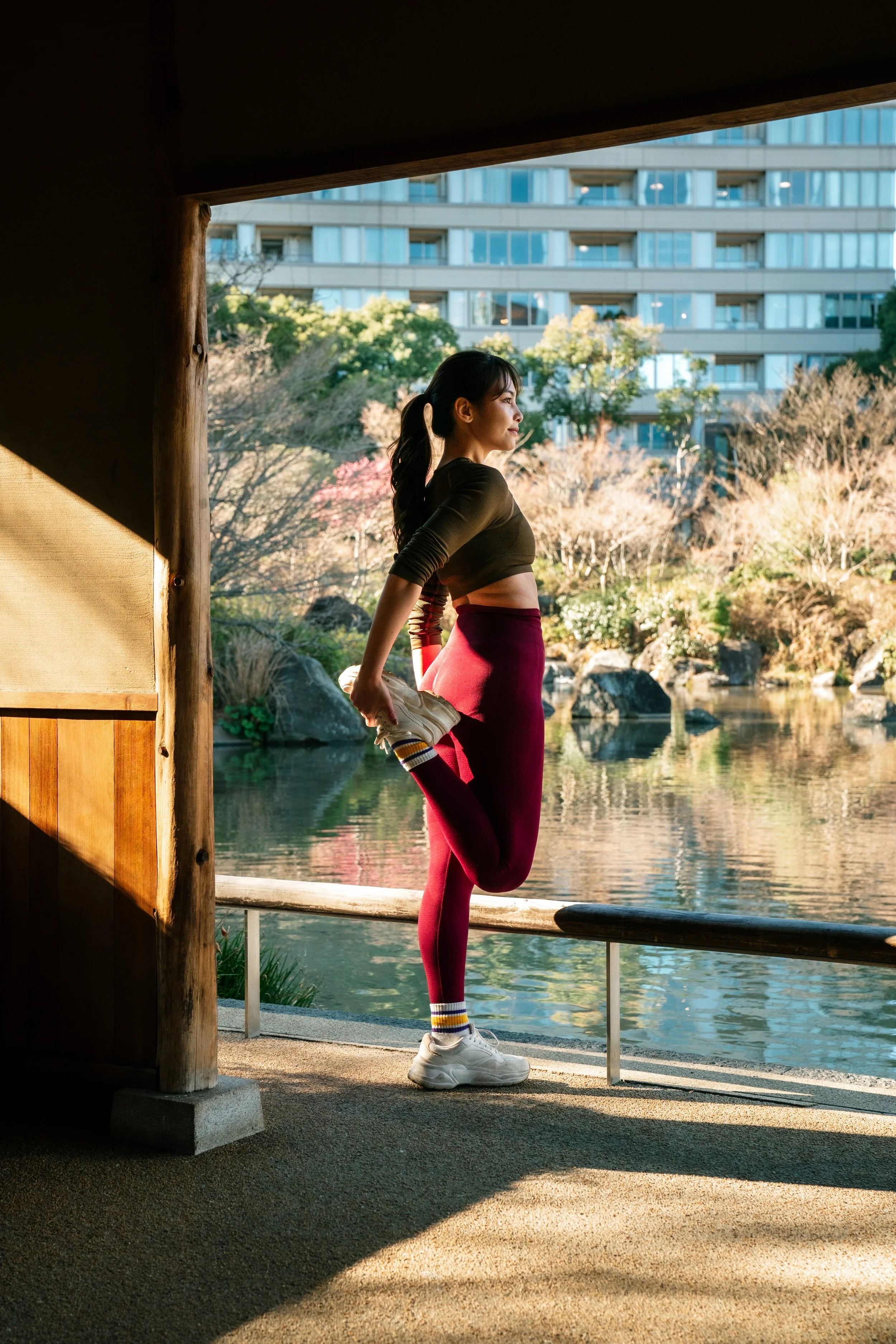 A woman stretching outdoors by a lake, holding her foot behind her and balancing on one leg, with buildings and trees in the background.
