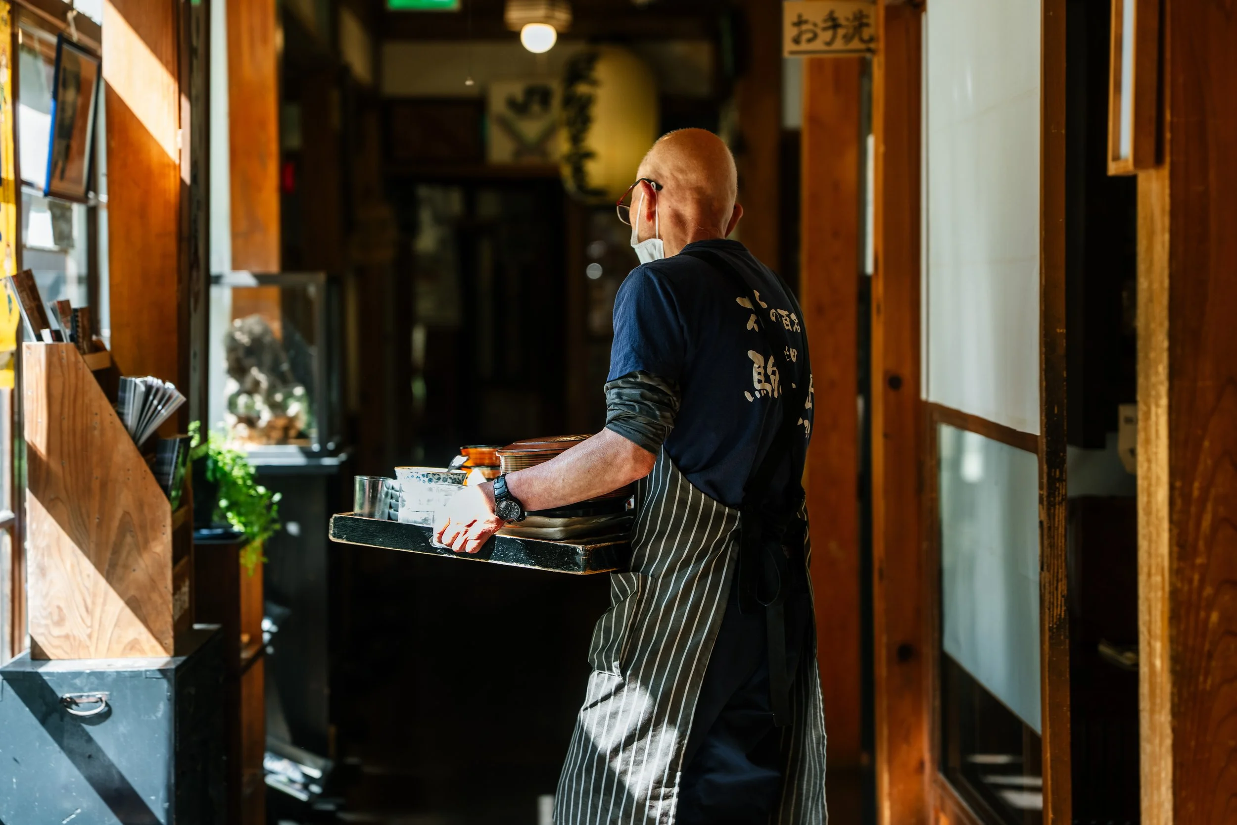 A person with a shaved head wearing a face mask, black shirt with white characters, and an apron, carrying a tray with glasses inside a wooden-framed restaurant or shop.