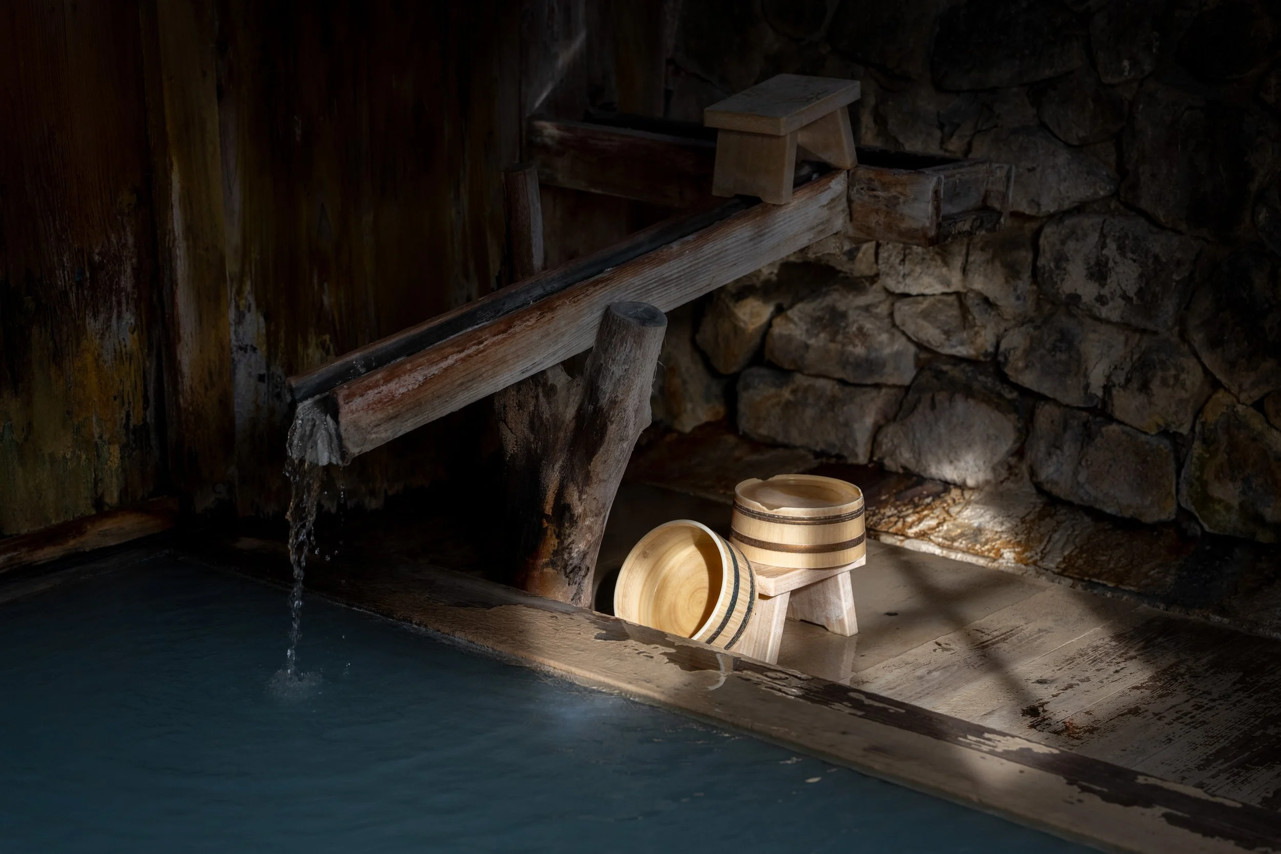 Wooden water trough with flowing water, two wooden cups and a small stool against a stone and wood wall.