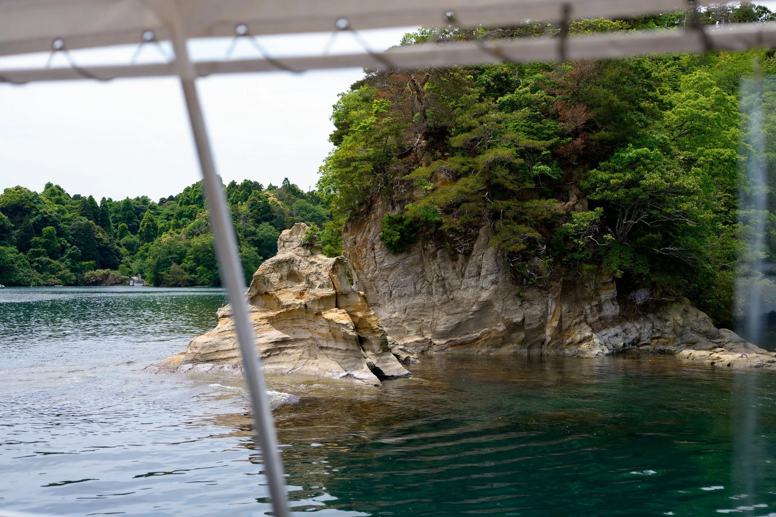 View of a rocky shoreline with green trees and a boat visible in the distance, seen from a boat or watercraft, with part of the boat's structure in the foreground. Noto, Ishikawa, editorial travel photography, Japan.