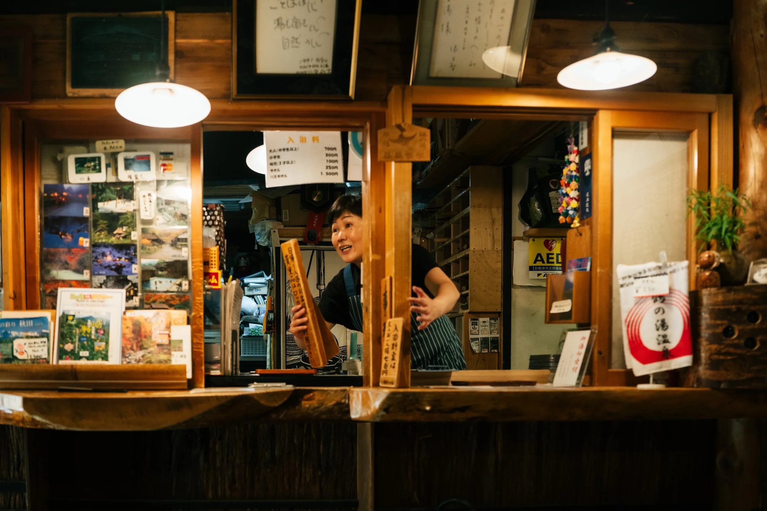 A woman smiling behind a wooden counter in a Japanese store, holding a wooden sign. The store has wooden walls, framed writings, photographs, and various signs and products displayed.