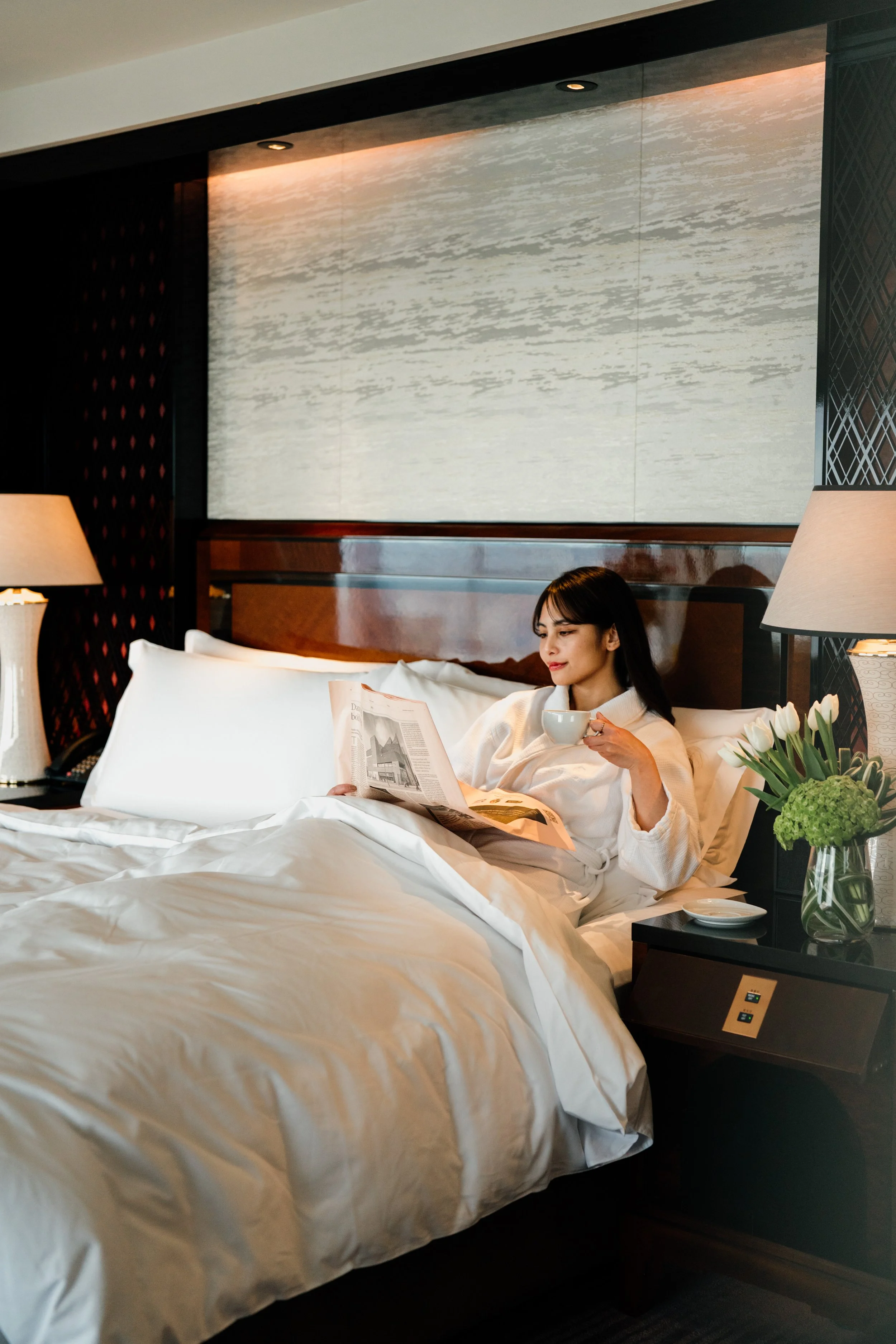 A woman sitting in bed in a hotel room, reading a newspaper while holding a cup. The room has modern decor with a large white bed, lamps, and a vase of tulips on a side table.