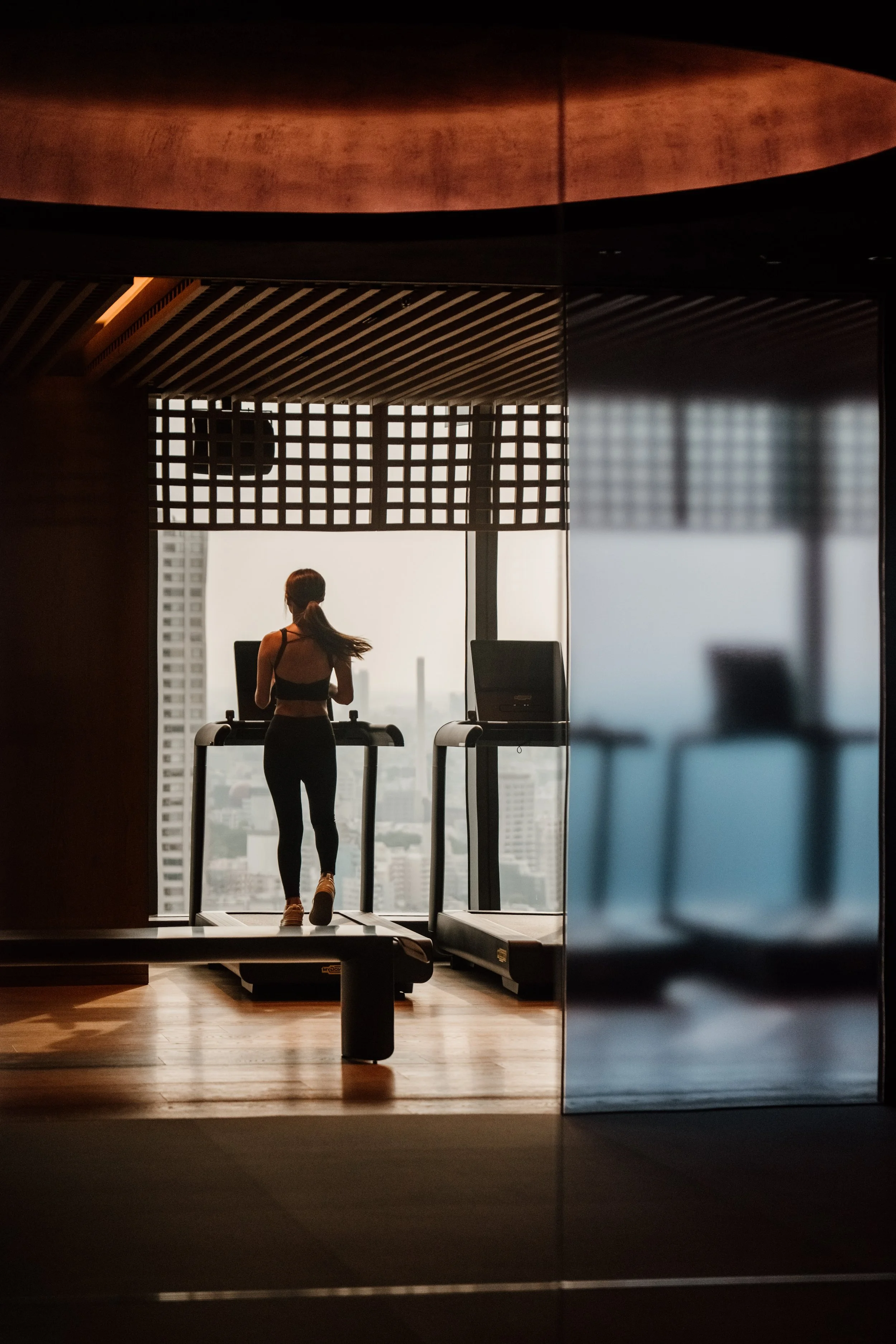 A woman using a treadmill in a modern gym with large windows showing a cityscape in the background at JW Marriott Tokyo hotel, editorial hospitality photography, Japan.