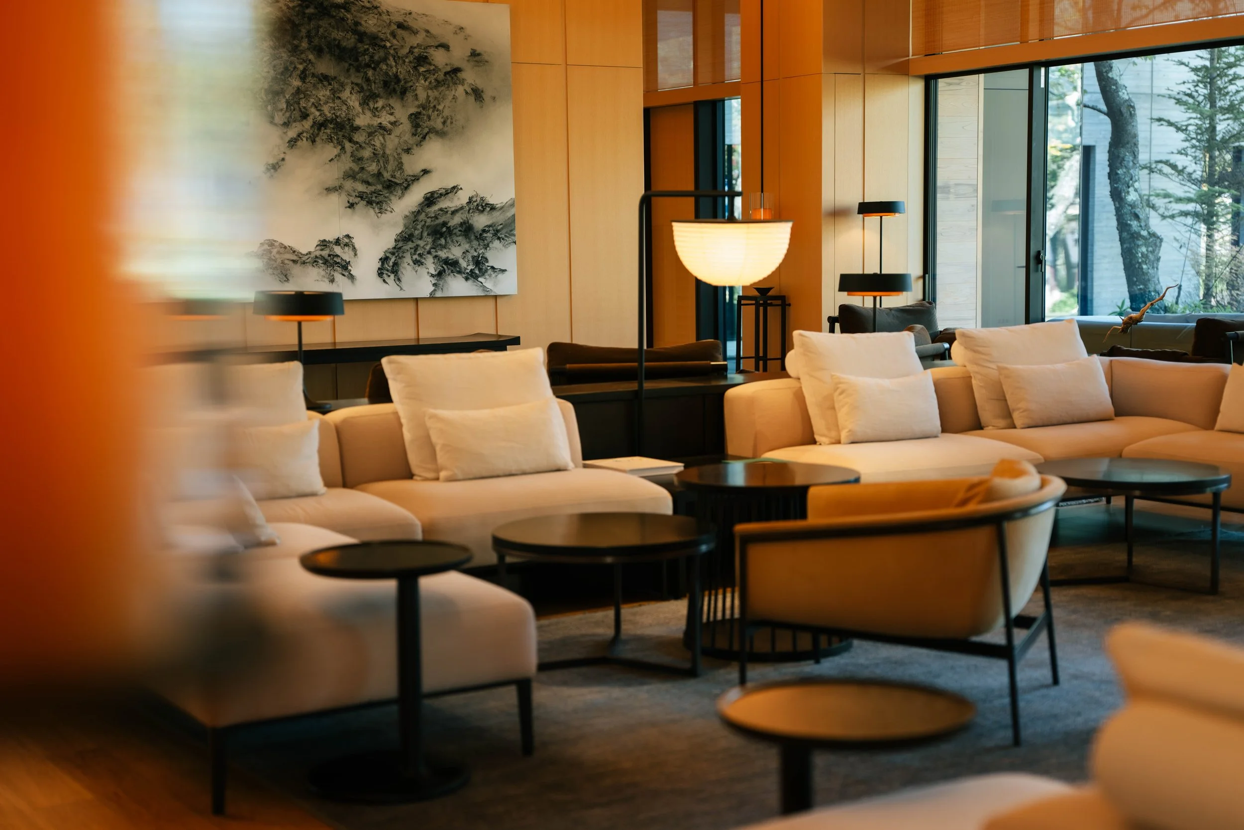 Modern living room with beige sofas, black side tables, a wooden wall, and large windows showing trees outside. There are black and orange lamps, a large painting with black ink strokes on a white background on the wall.