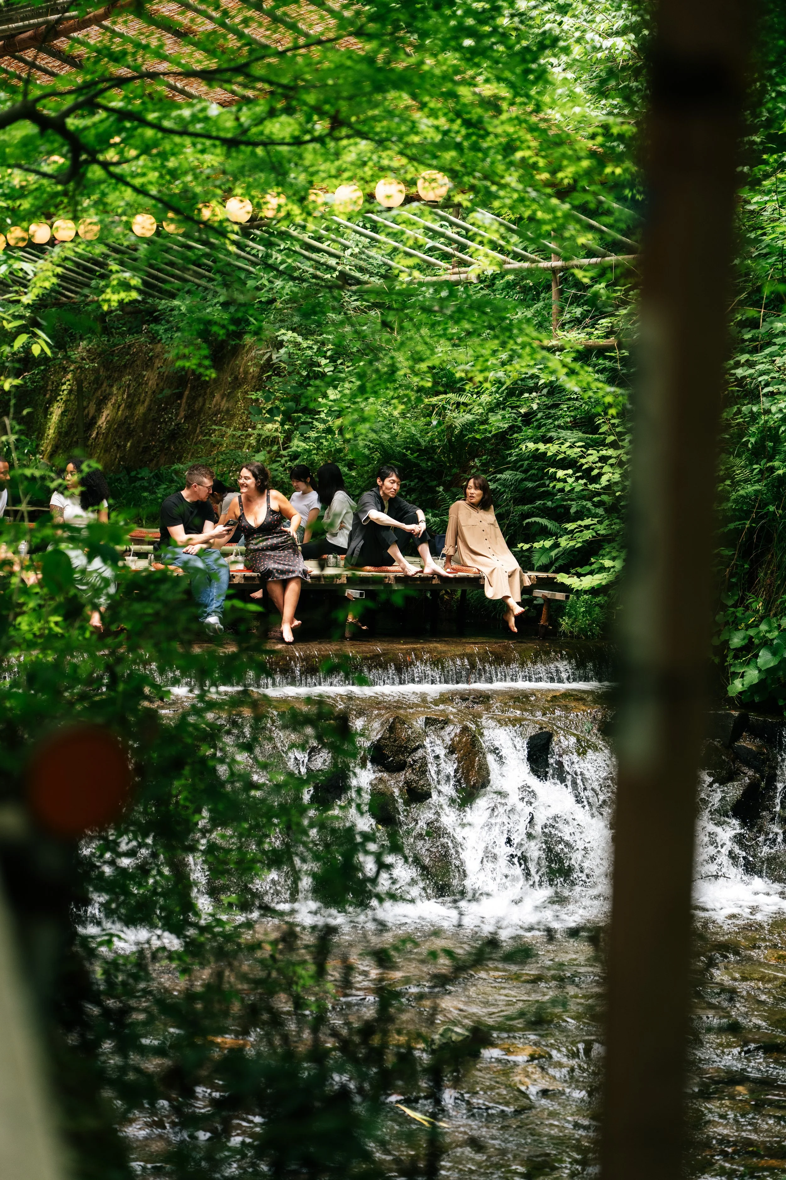 People sitting on a wooden platform near a flowing stream in a lush forest, with trees and green foliage. Lanterns are hanging above them, and some are engaged in conversation.