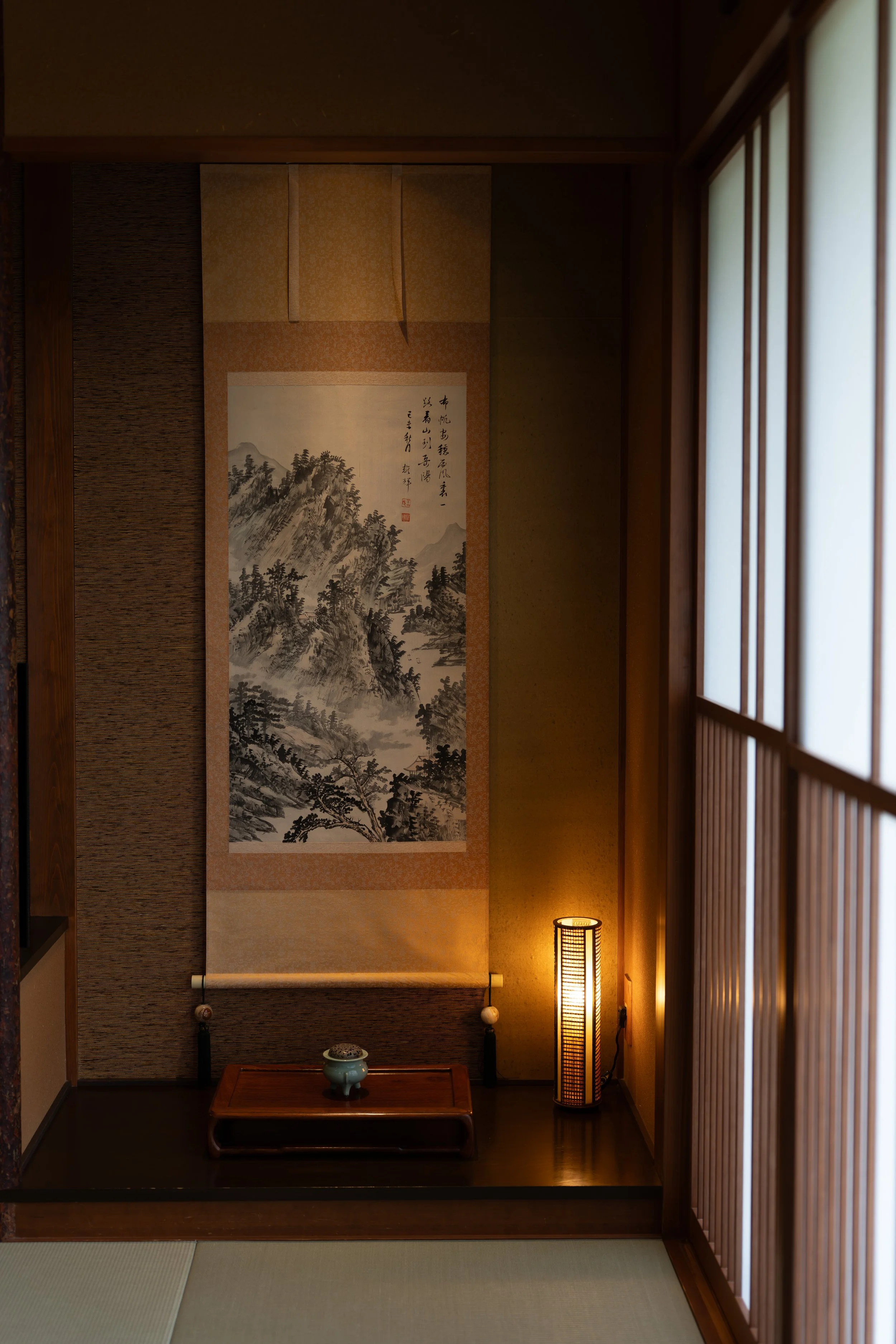 Traditional Japanese interior room with shoji sliding doors, a hanging painting of mountainous landscape, and a small wooden table with a ceramic tea bowl. A tall, cylindrical paper lantern is in the corner, and tatami mats are on the floor.
