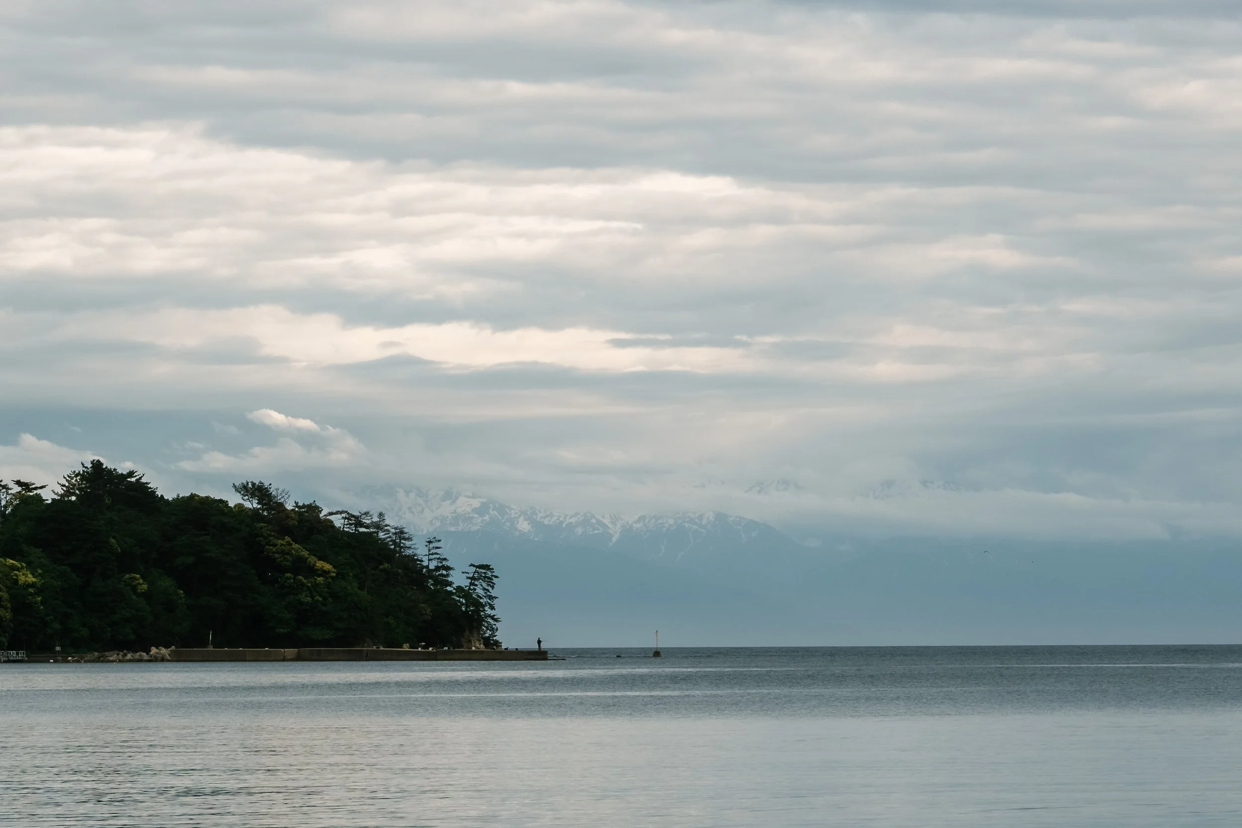 A view of Tsukumo bay with a hill on the left, a cloudy sky, snow-capped mountains in the background, and a small pier with a person standing at the end, near the water. Noto, Ishikawa, editorial travel photography, Japan.