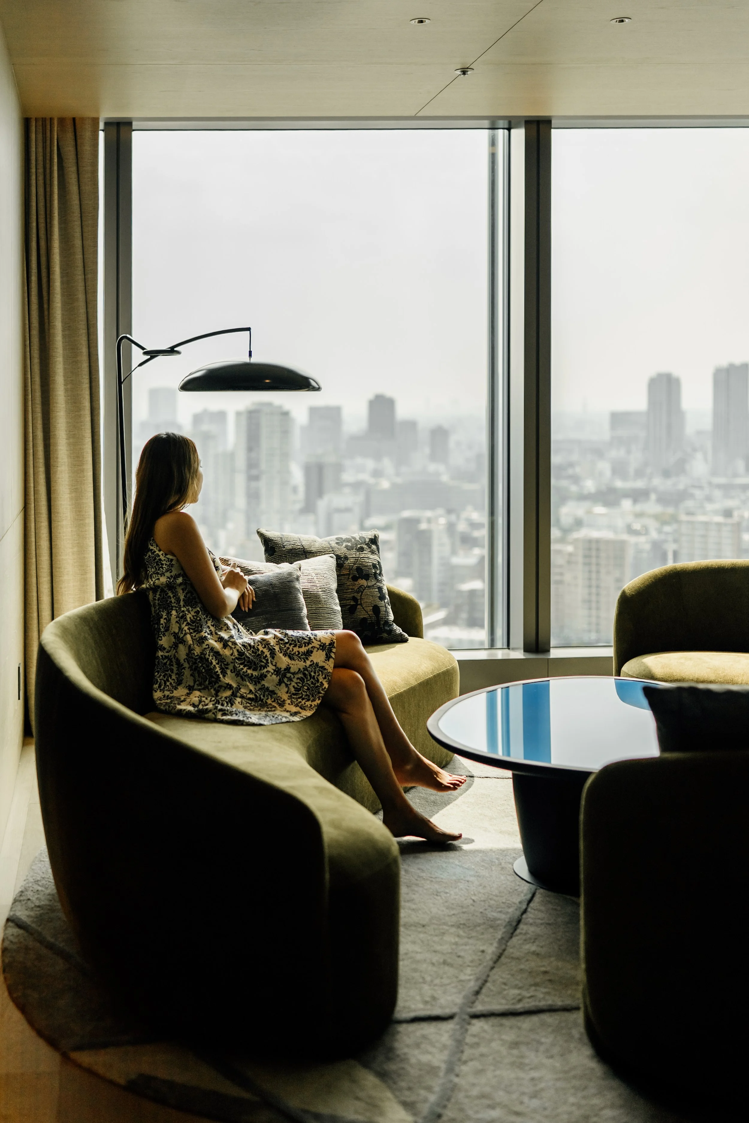 A woman with long hair sitting on a green sofa in a modern living room, looking out a large window with a view of a city skyline at JW Marriott Tokyo hotel, editorial hotel photography, Japan.