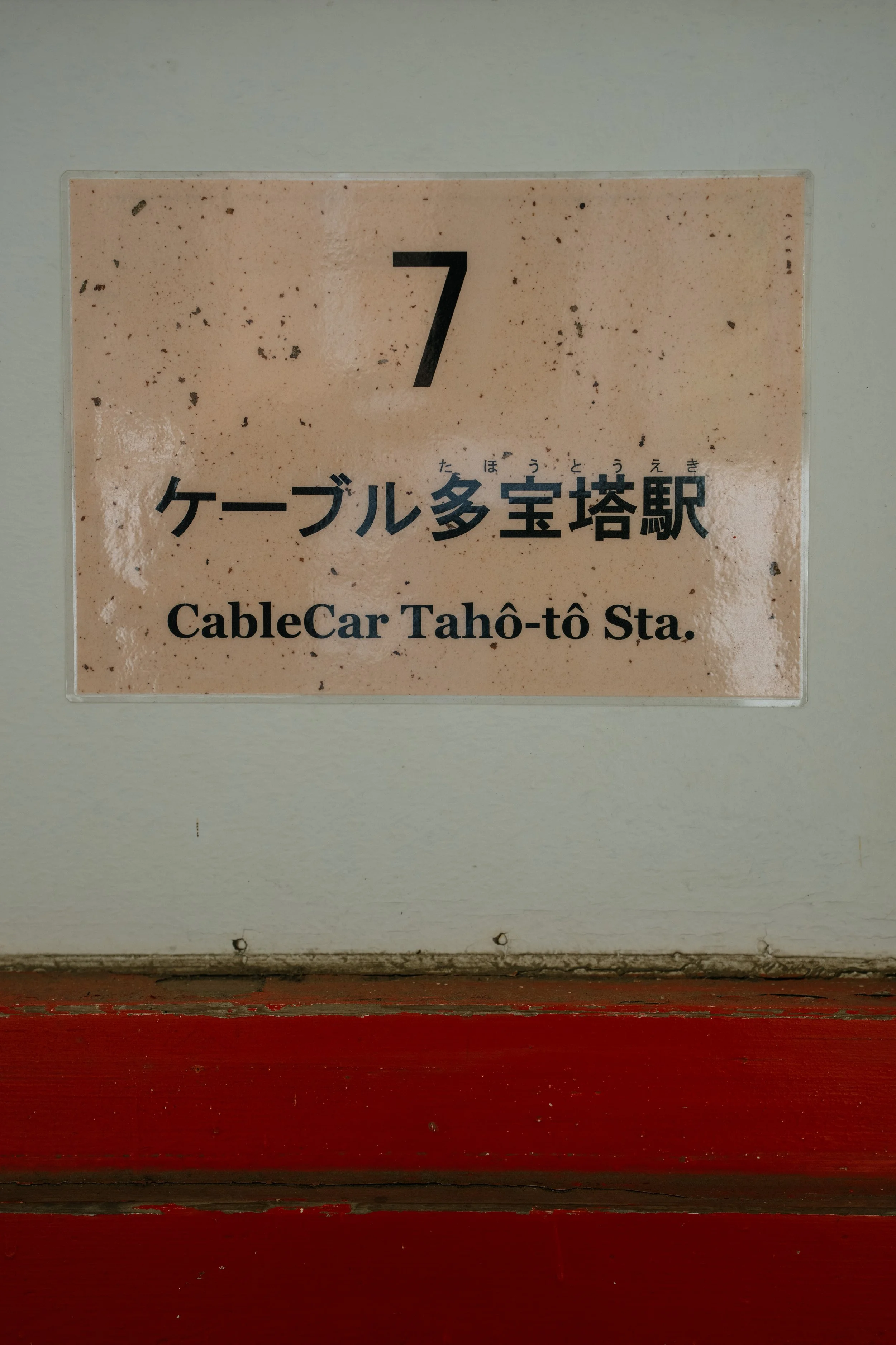 A sign with the number 7, Japanese characters, and English text reading 'CableCar Taho-tô Sta.' mounted on a wall above a red painted surface.