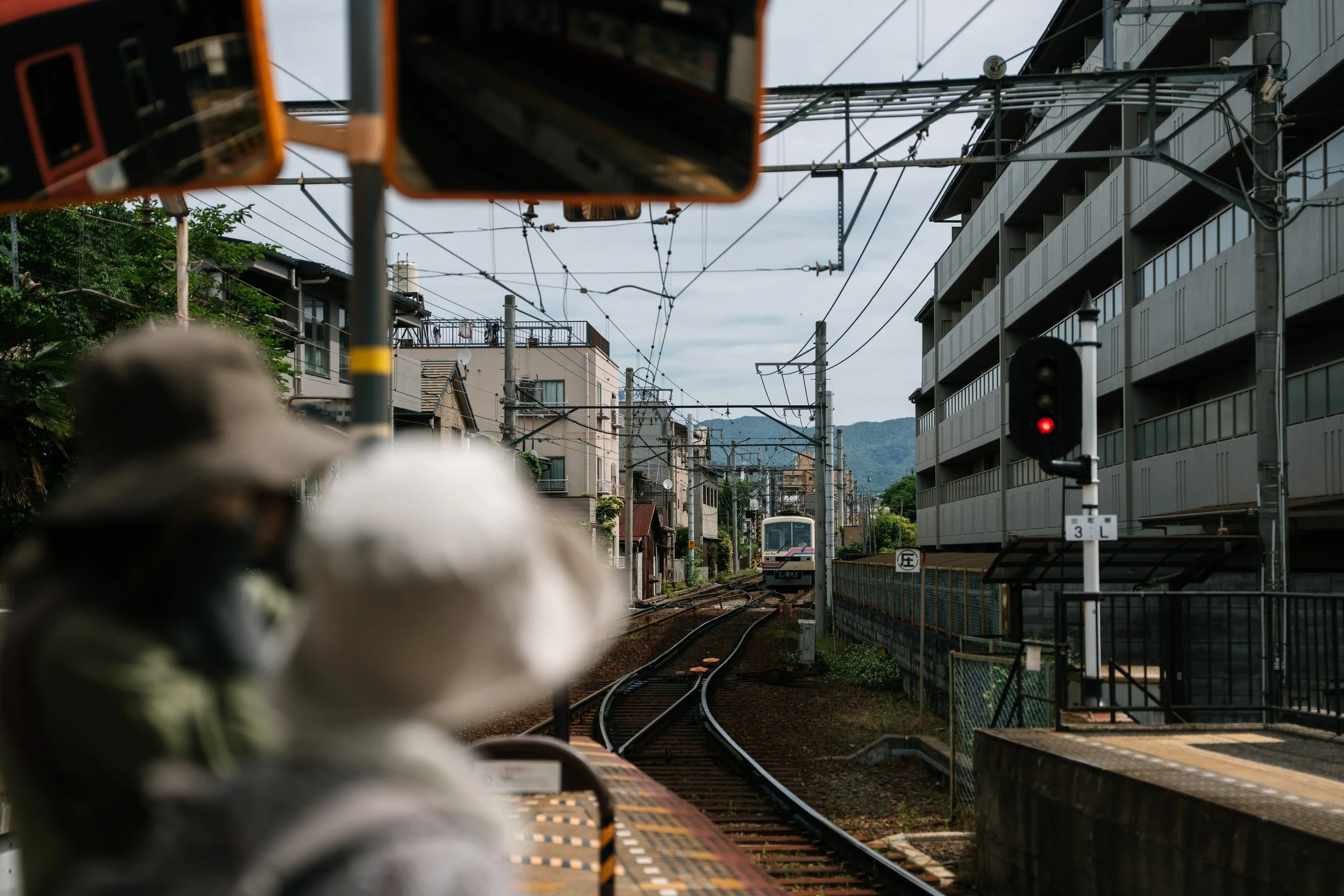 View from a train platform showing railway tracks, a passing train, overhead electrical wires, and urban buildings, with a person in the foreground.