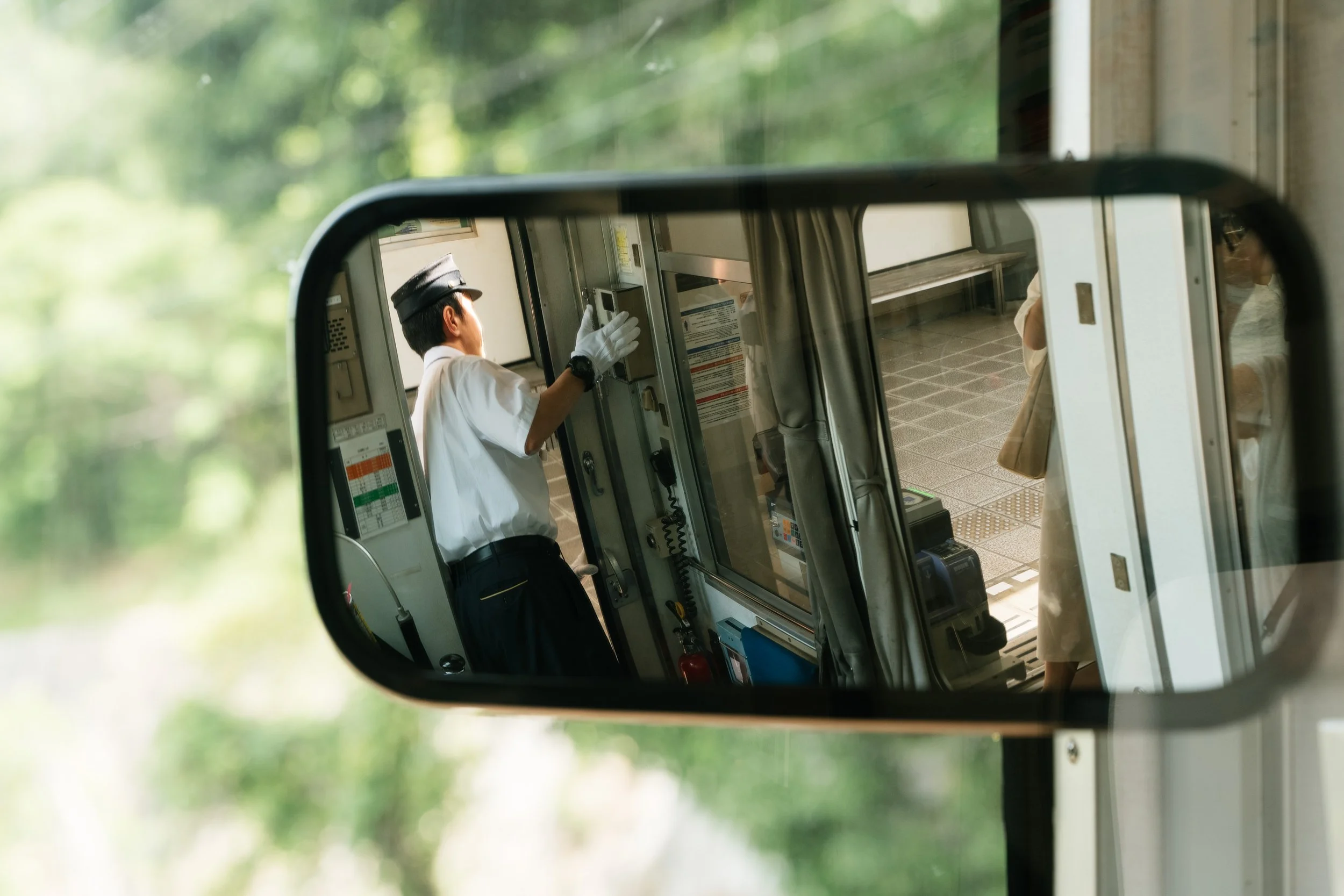 Reflections of a train conductor and passengers seen in a train station mirror with trees outside.