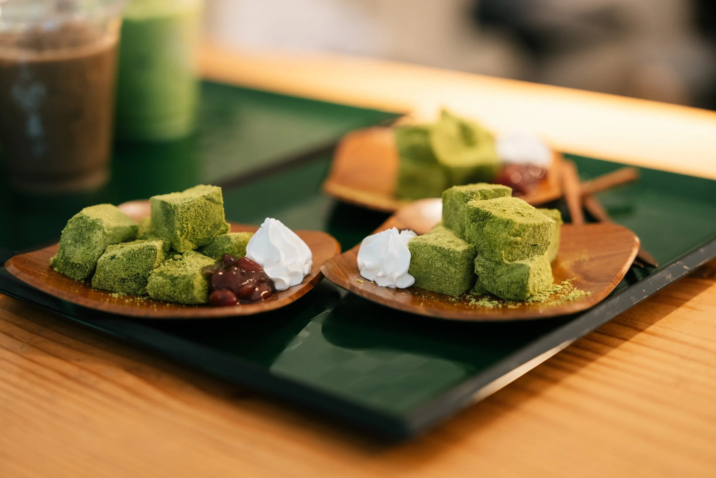 Matcha green tea flavored desserts on a wooden platter, including green tea cake pieces, whipped cream, and red bean paste, with another dessert and drinks in the background.