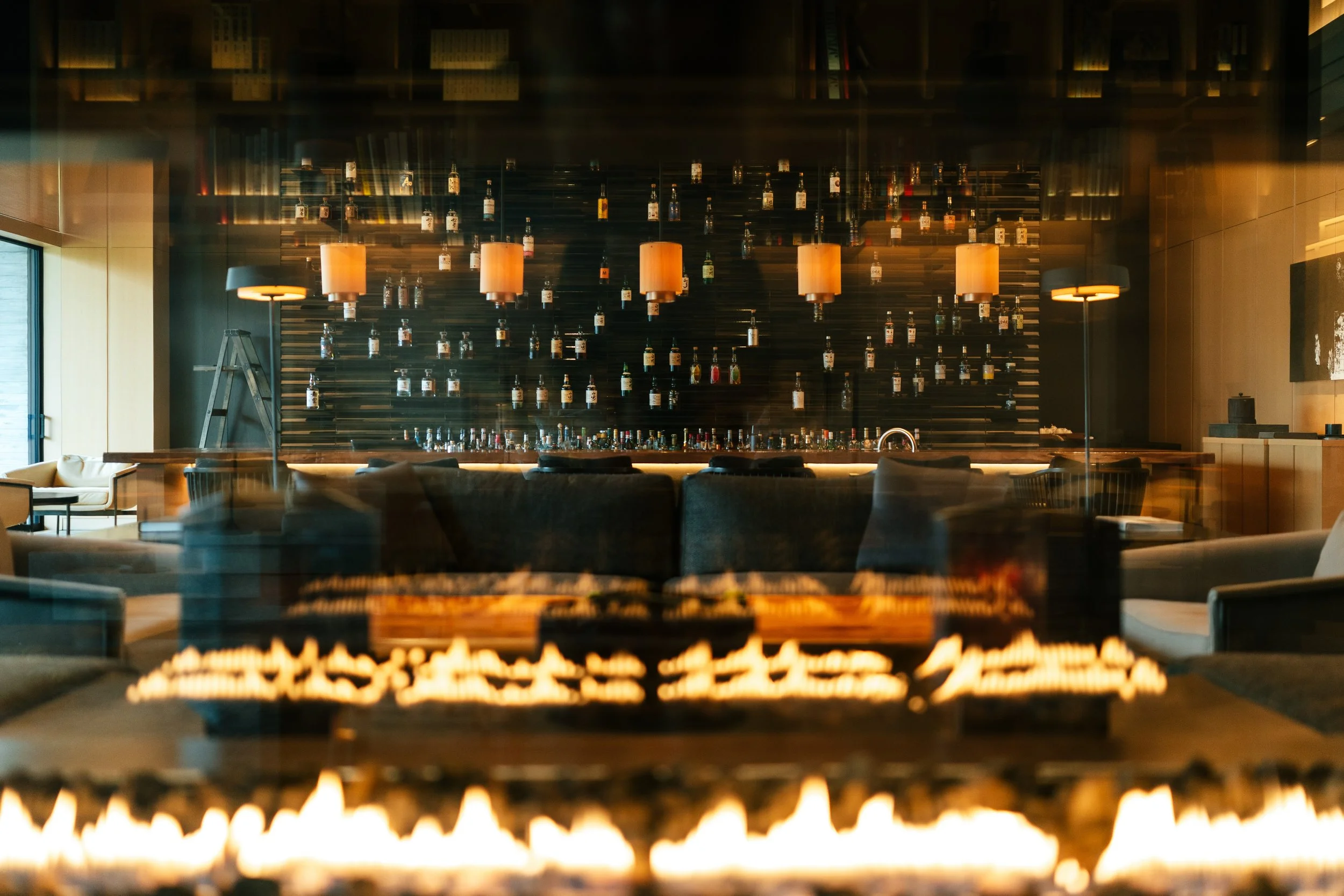 Interior of a modern bar or lounge with warm lighting, hanging pendant lamps, a wall-mounted shelf with bottles, seating area with couches, and a stone fire pit in the foreground.