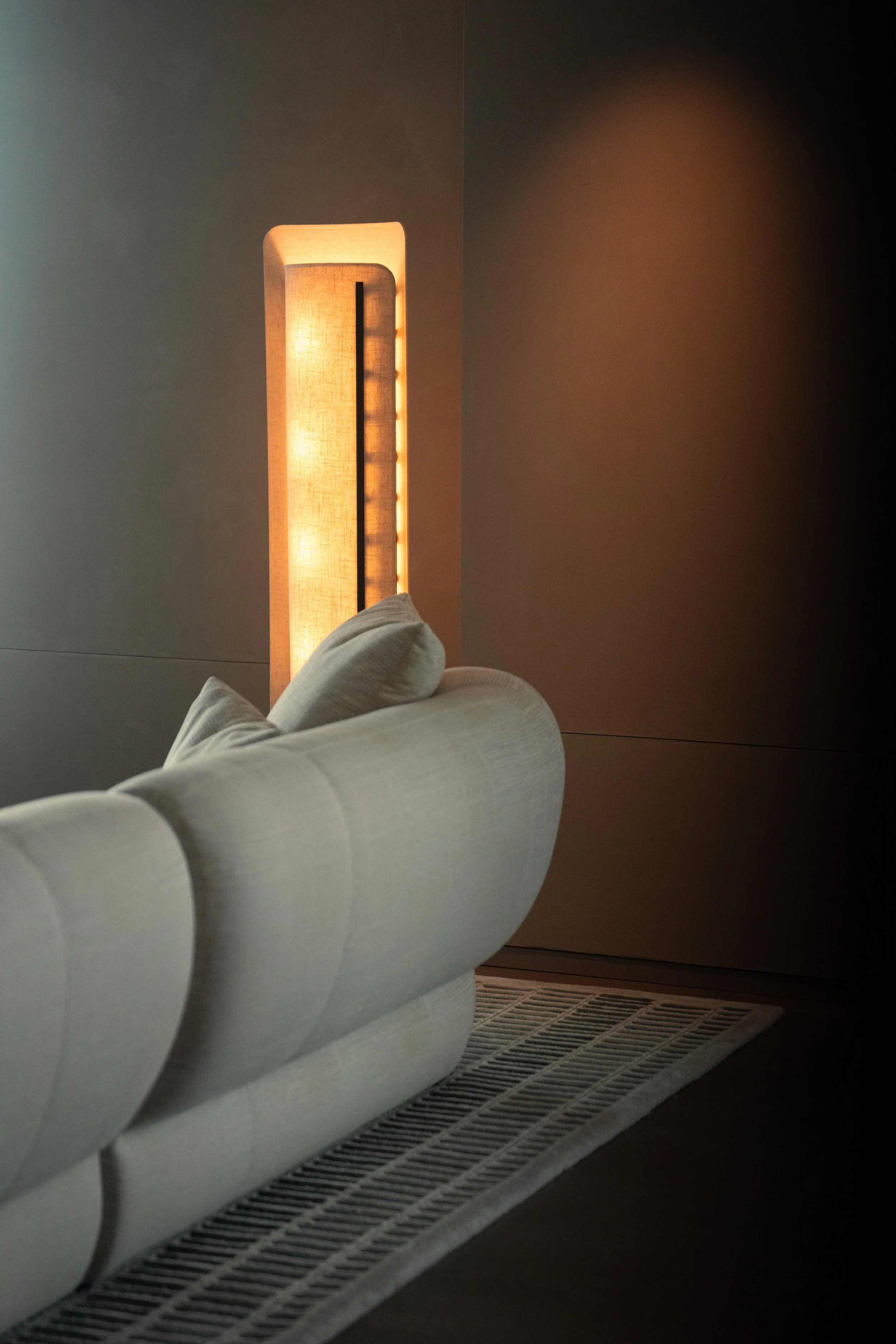 A contemporary bedroom nightstand lamp with a fabric shade and a warm glow, positioned next to a white upholstered bed and a black and white striped rug on a hardwood floor.