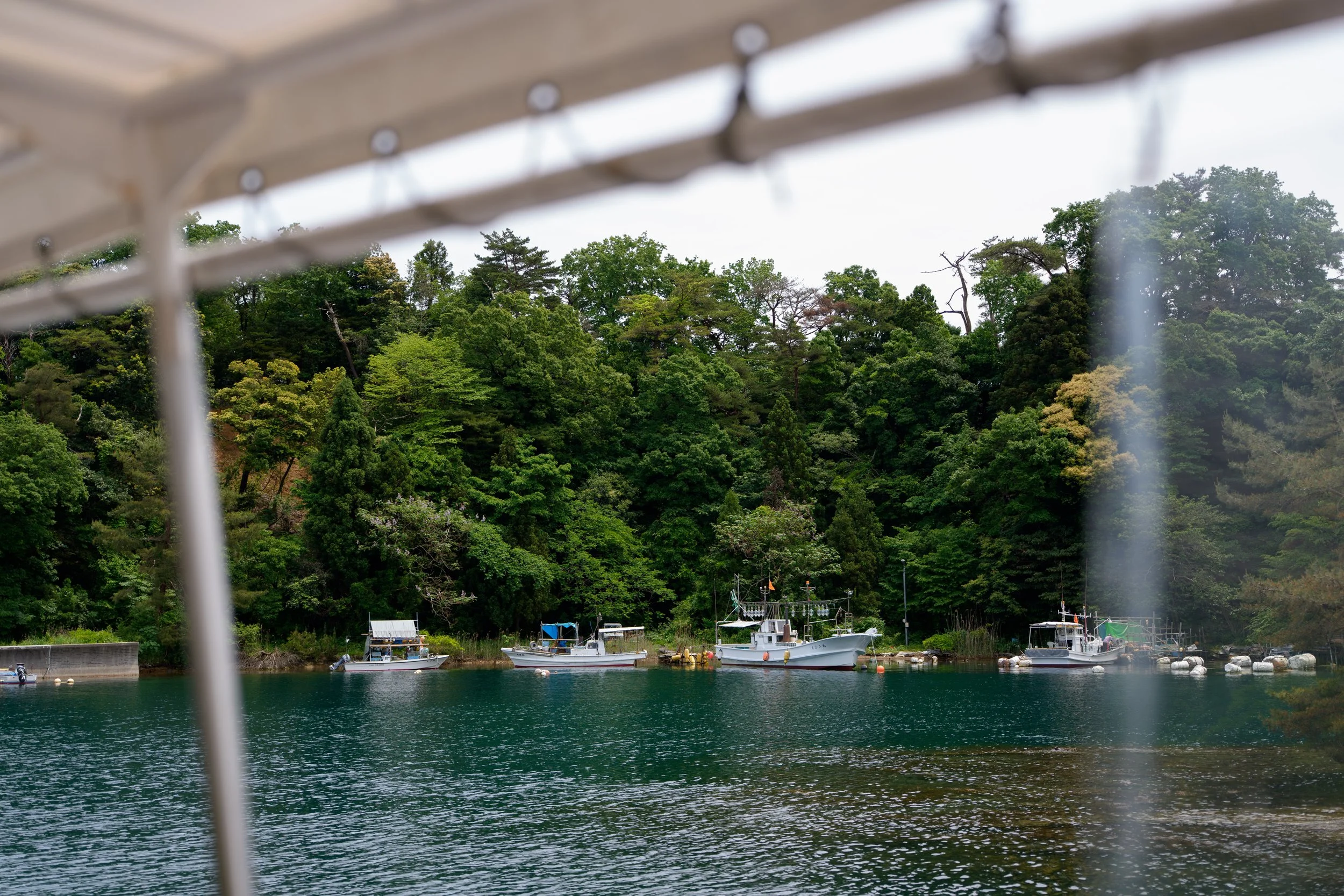 View of a small harbour with boats docked in Tsukumo bay and lush green trees in the background, seen through the frame of a boat or dock. Noto, Ishikawa, editorial travel photography, Japan.