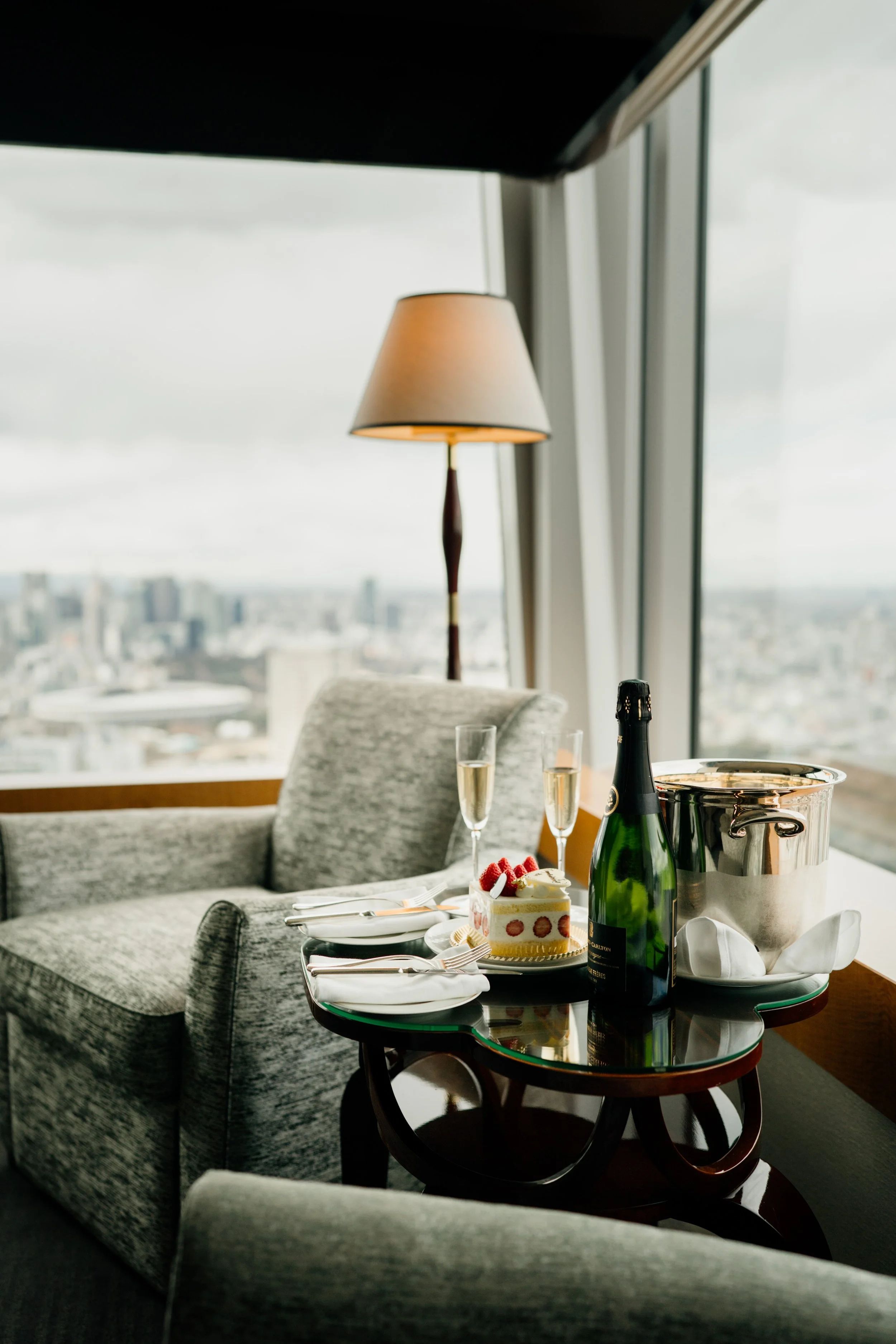 A dining table with a dessert cake, two glasses of champagne, a champagne bottle, and an ice bucket, in front of large windows with a city view at The Ritz-Carlton, Tokyo. Editorial hospitality photography, Japan.