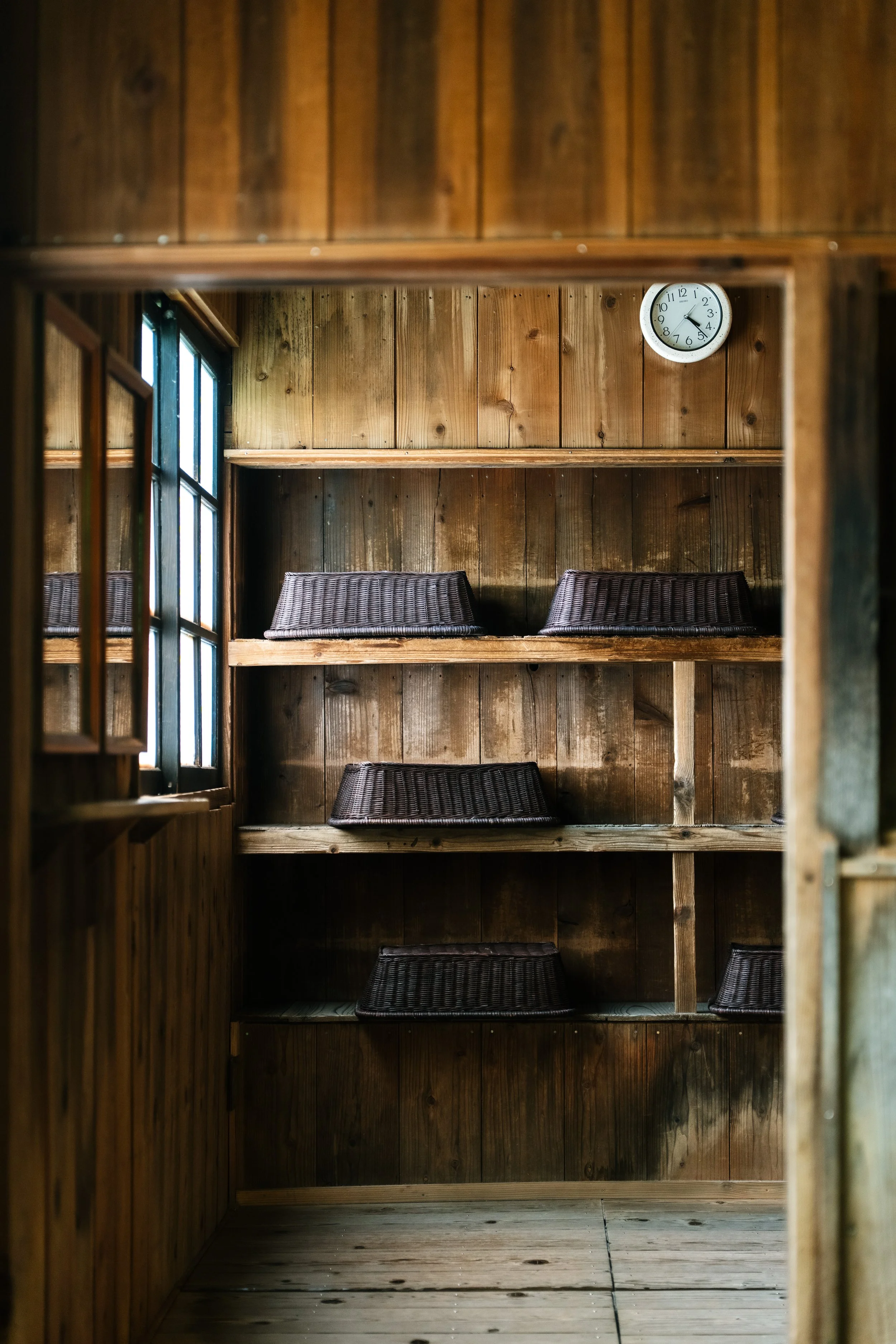 Wooden wall with shelves and a clock showing approximately 3:15. Wicker baskets are placed on the shelves, and window with sunlight coming in is visible.