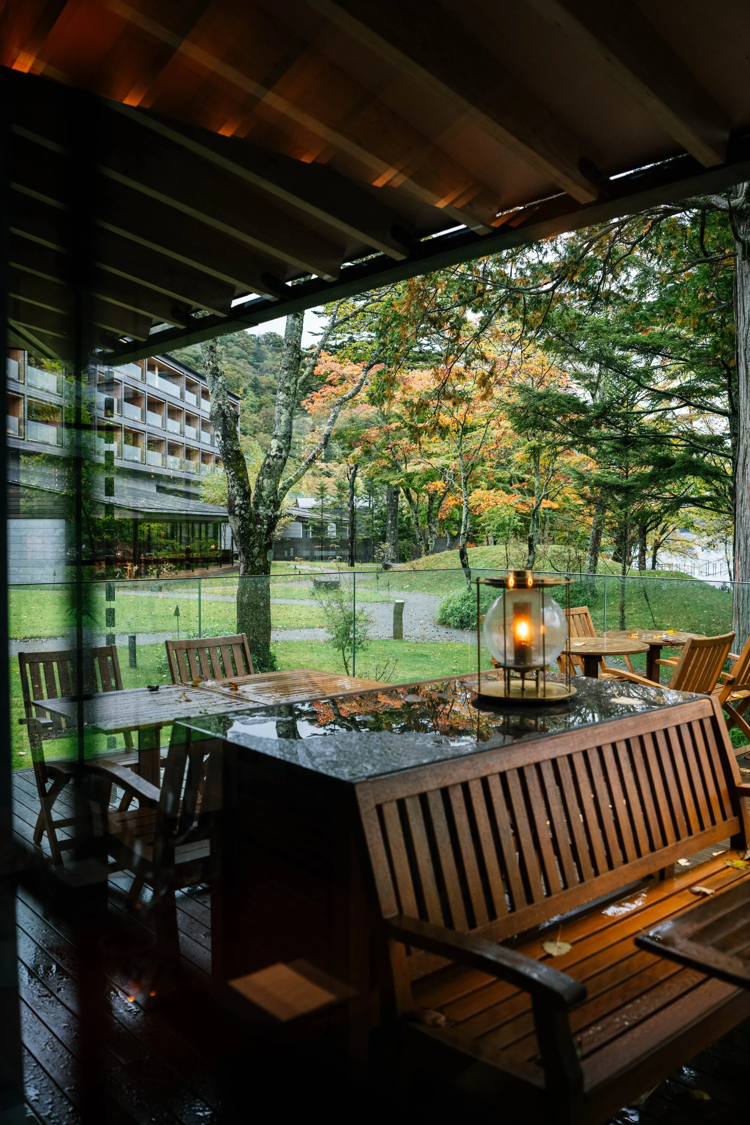 Outside view from a covered deck showing a wooden table and chairs, a lantern with a lit candle, green trees with colorful autumn leaves, and a modern apartment building in the background on a rainy day.