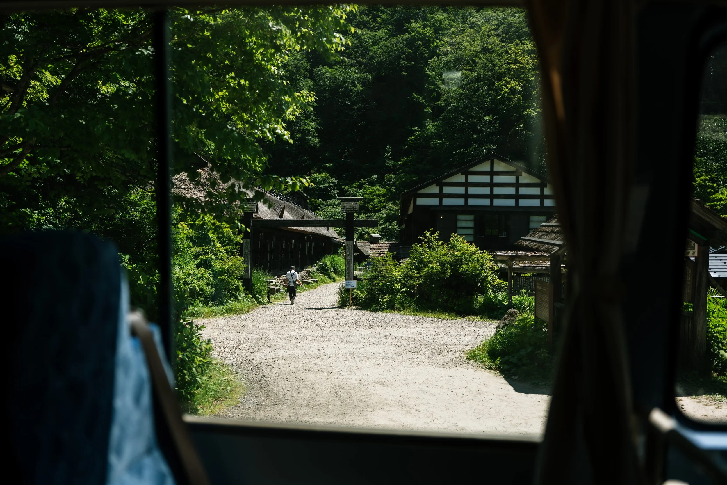 View of a rural pathway with a person walking, houses, and lush green trees in a mountainous area, seen from inside a vehicle.