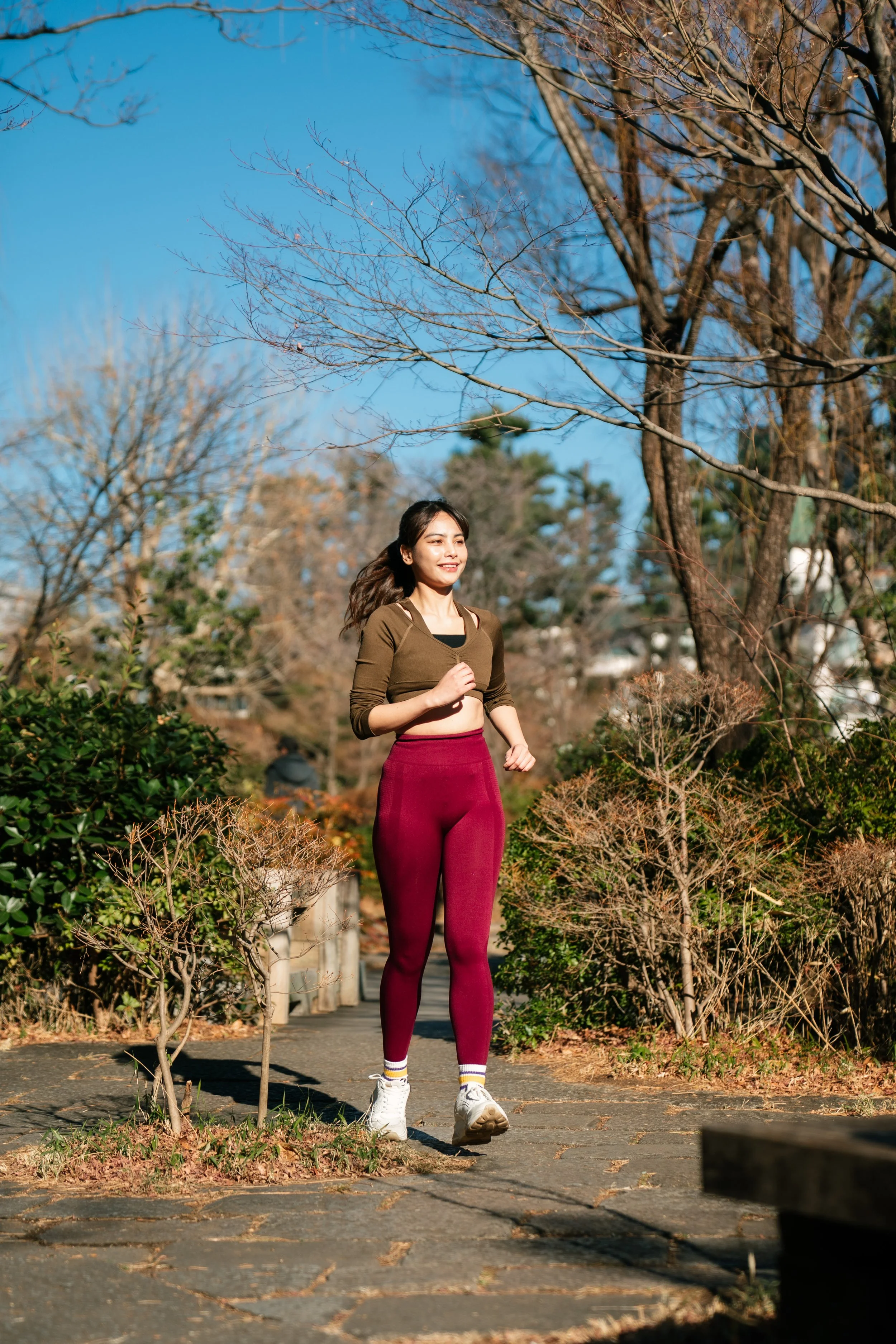 A woman jogging on a pathway in a park with leafless trees and bushes around, wearing athletic clothing including maroon leggings, a brown top, and white sneakers.