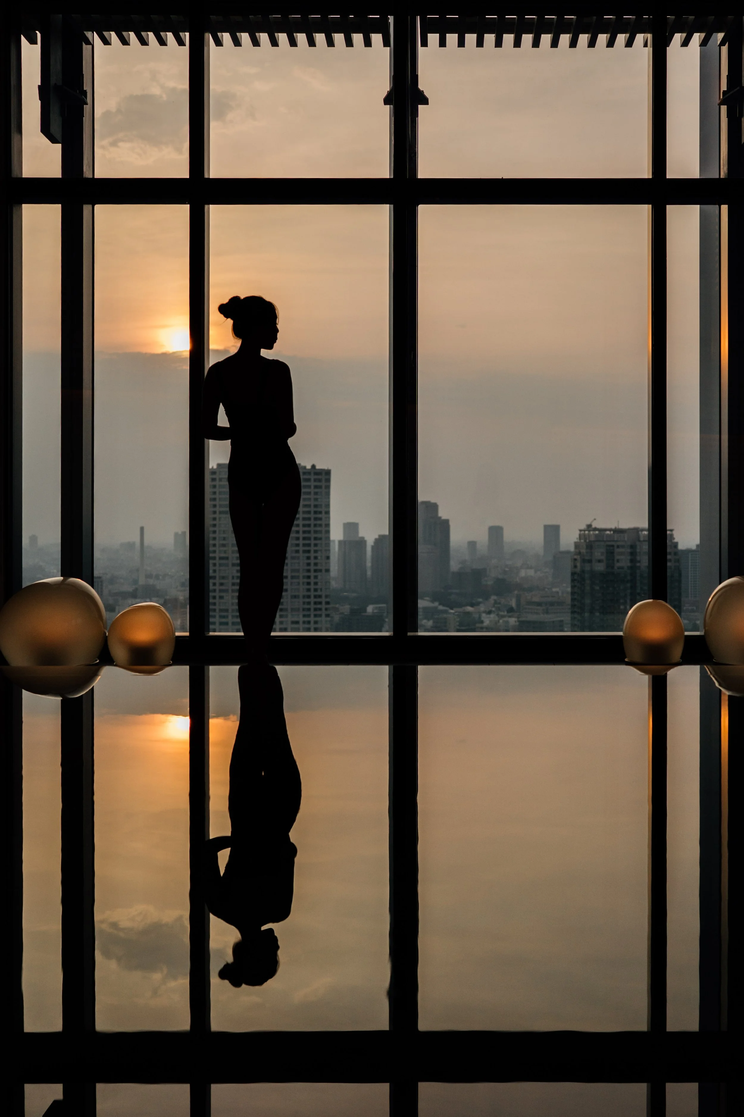 Silhouette of a woman standing in front of a large window during sunset, with city buildings in the background and a reflection on the swimming pool at JW Marriott Tokyo hotel, editorial hospitality photography, Japan.