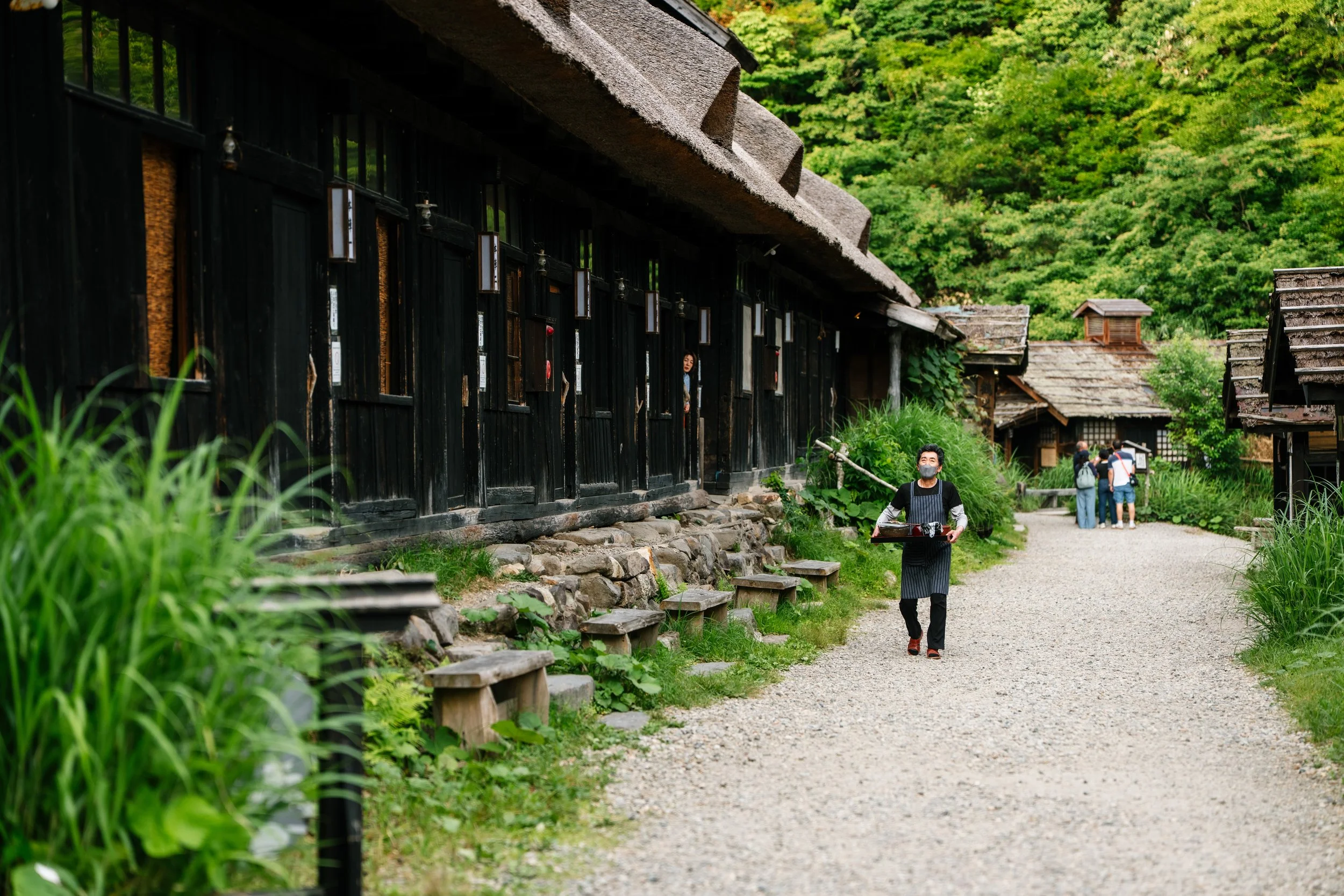 A person wearing a face mask and striped apron walking on a gravel pathway in a traditional Japanese village with black wooden buildings and lush green trees.